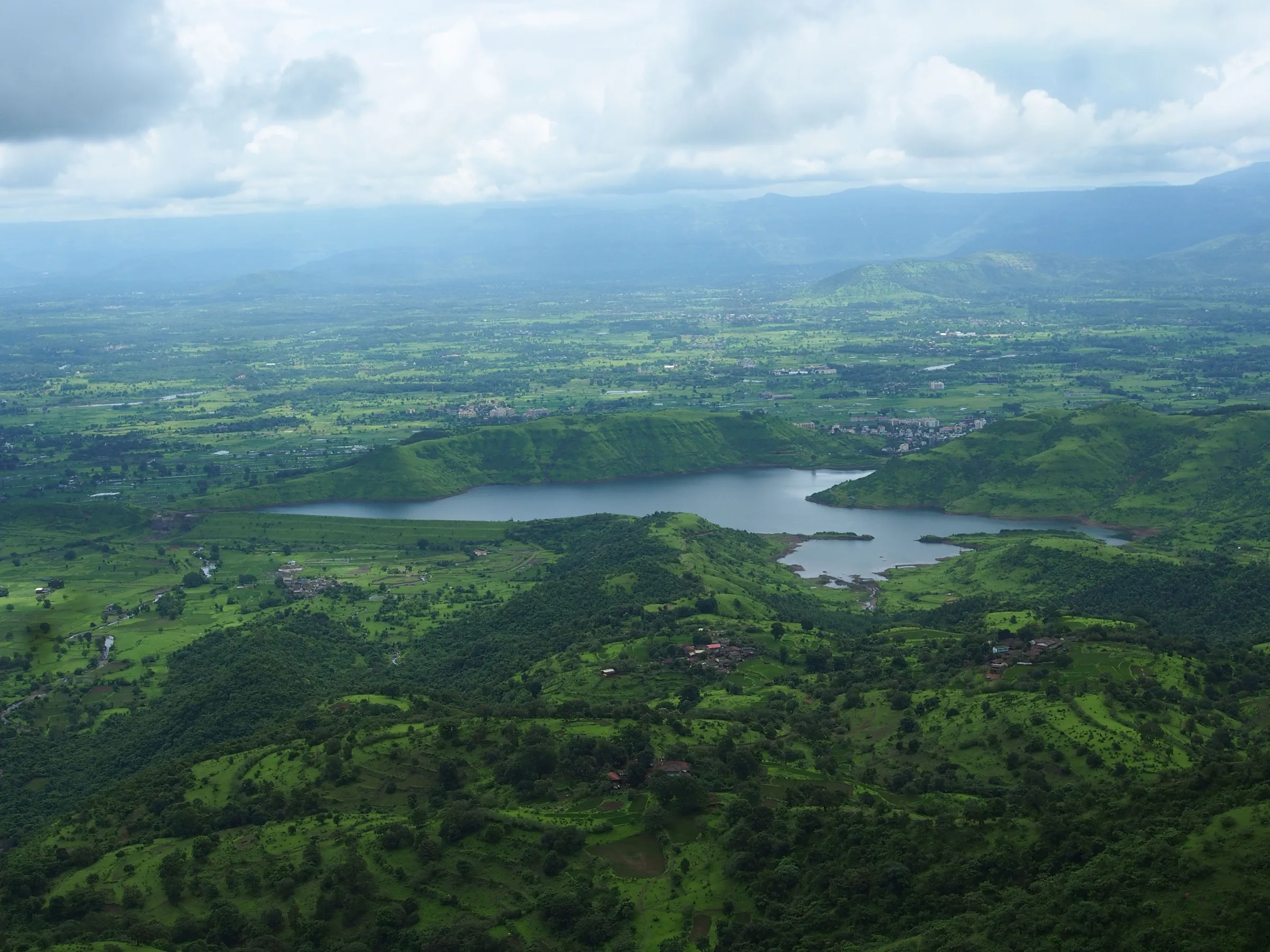 Dhom lake near Diksal seen from the green hills above, with farmland and the Karjat plain under monsoon clouds