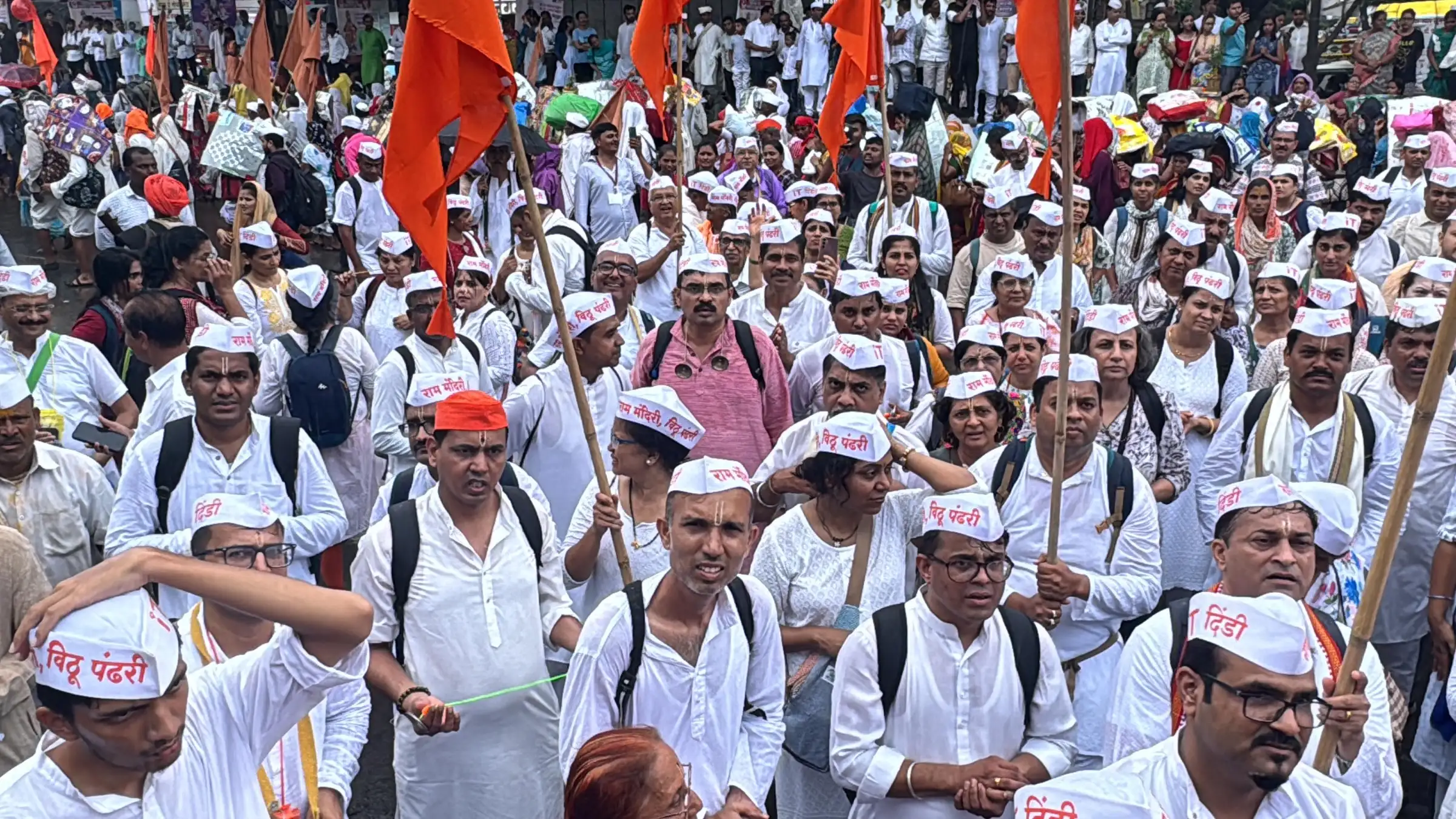 A dindi of warkaris in white caps reading Vithu Pandhari, with saffron flags, walking the Pune to Saswad stretch of the Wari