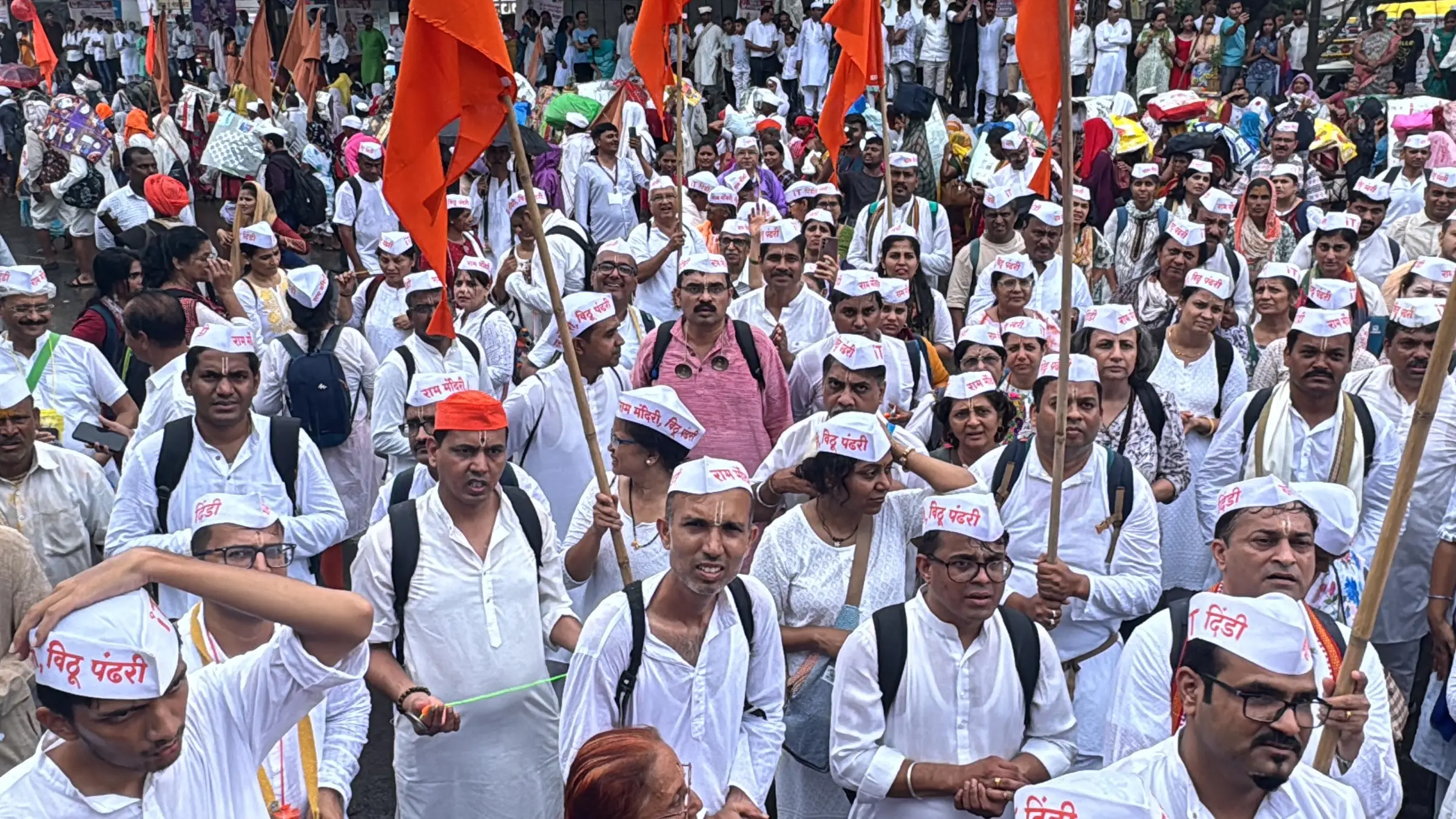 A dindi of warkaris in white caps reading Vithu Pandhari, with saffron flags, walking the Pune to Saswad stretch of the Wari