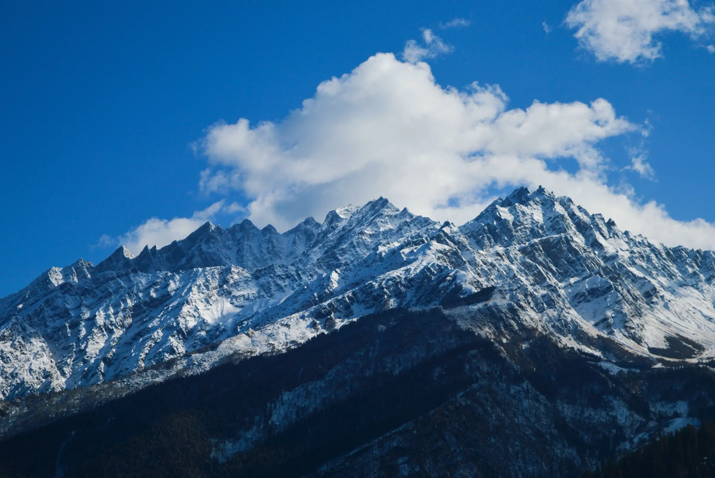 Jagged snow-dusted ridge above dark forested slopes under a blue sky with white clouds, near Dronagiri