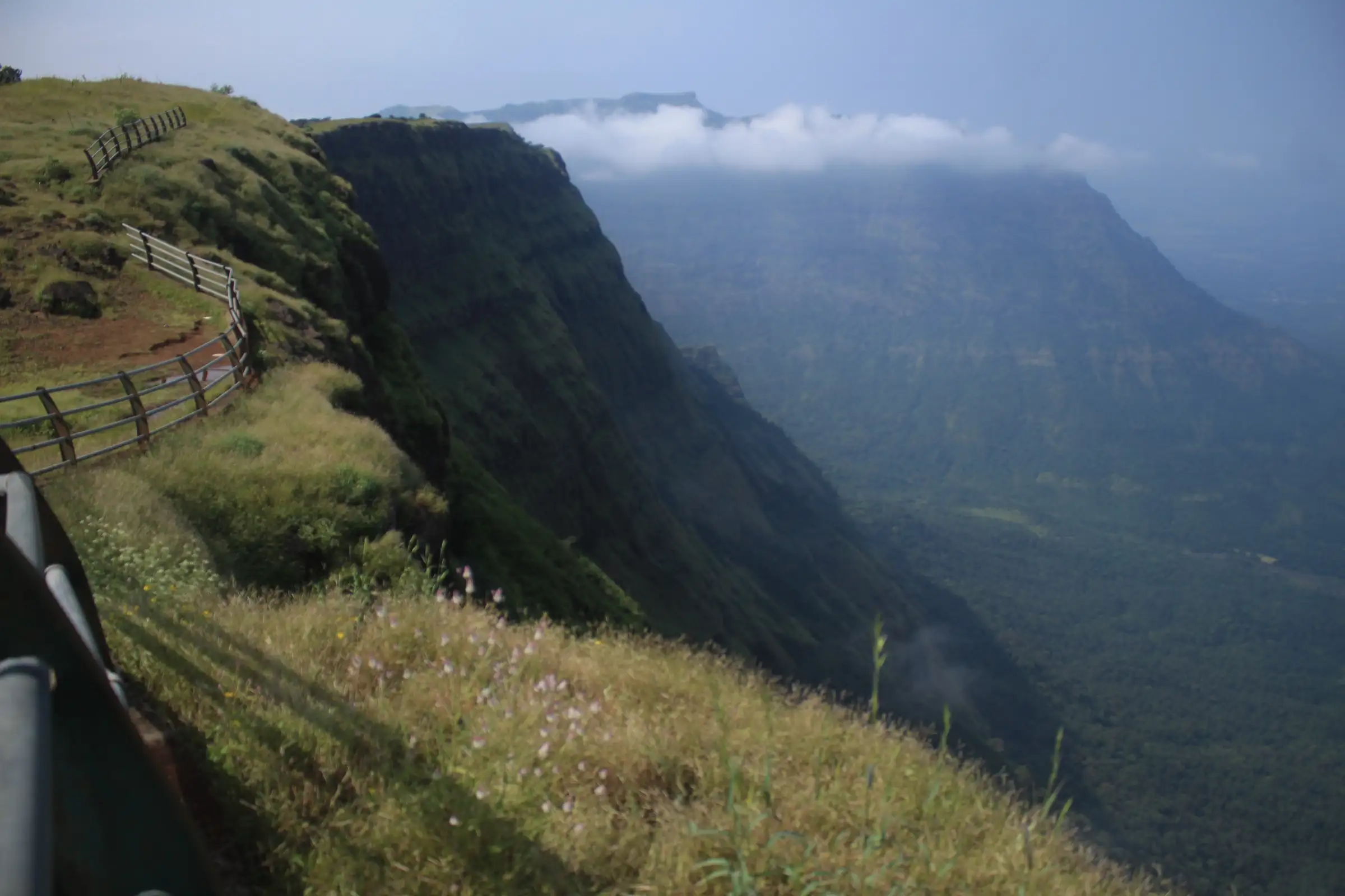 Grassy ridge with a wooden fence beside the sheer basalt cliff of the Sahyadri escarpment near Durg, with cloud over the flat-topped range beyond