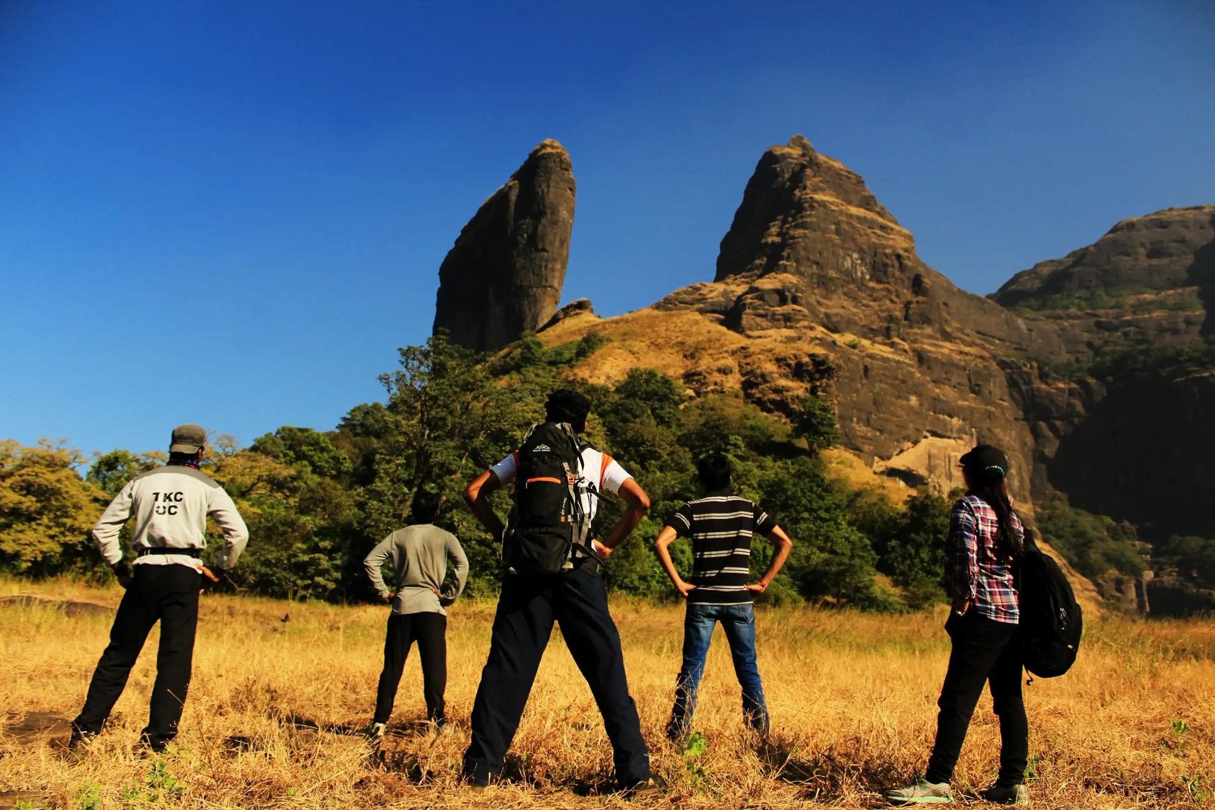 Trekkers standing in dry grass on the plateau looking up at the thin rock fin of Bhairavgad and the cliffs beside it