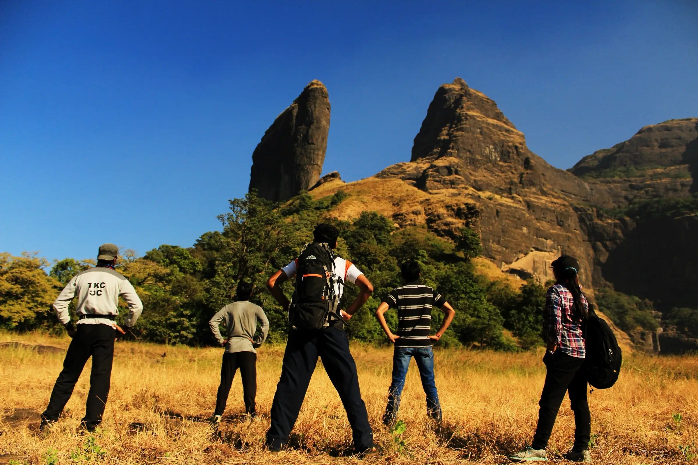 Trekkers standing in dry grass on the plateau looking up at the thin rock fin of Bhairavgad and the cliffs beside it