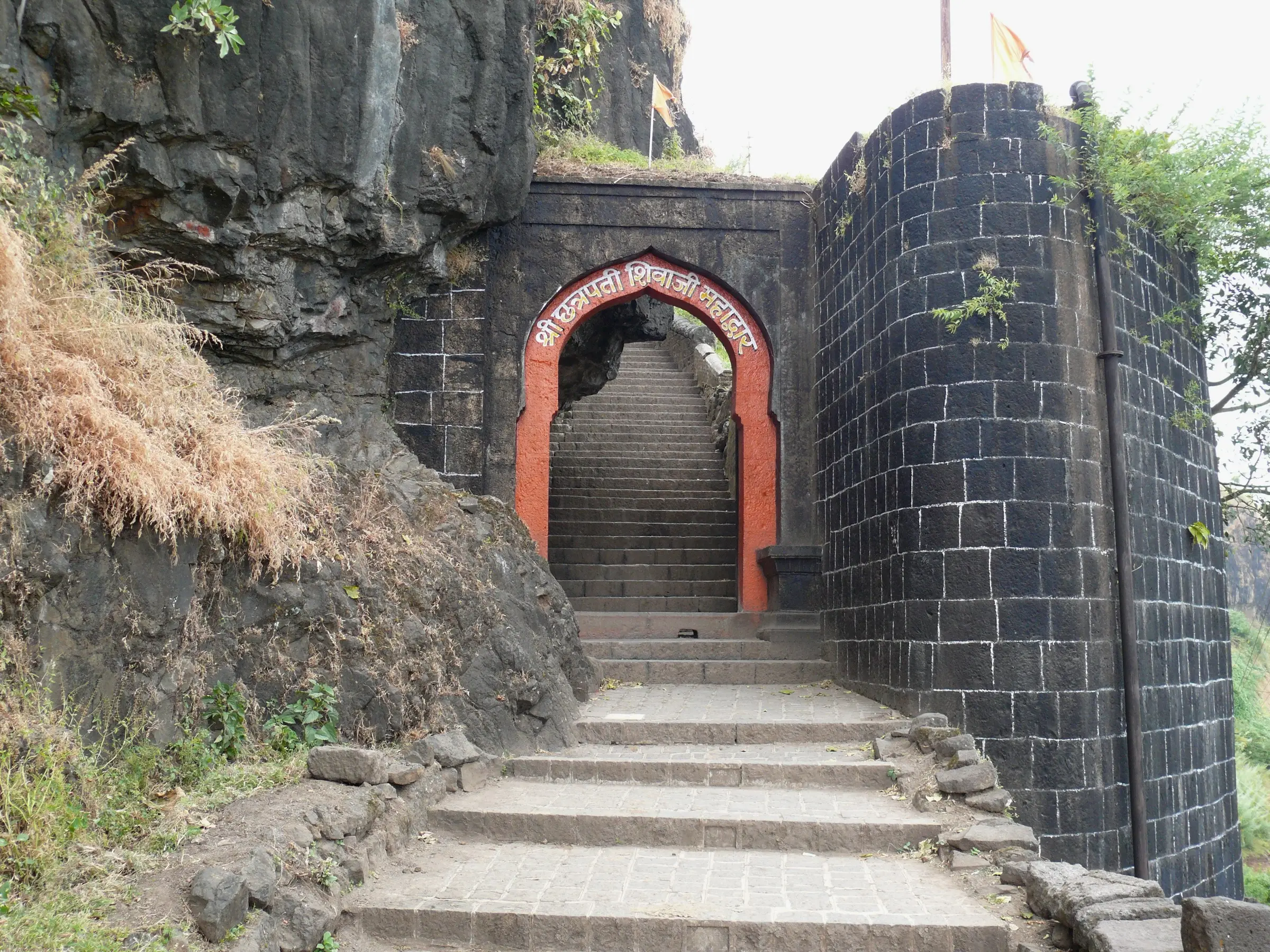 Stone steps climbing through the arched Shivaji Mahadarwaja gate of Sajjangad beside a round black-stone bastion