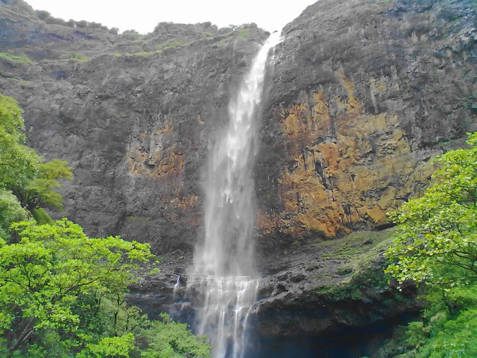 A tall thin waterfall dropping down a dark basalt cliff streaked with orange rock, with green trees in the foreground