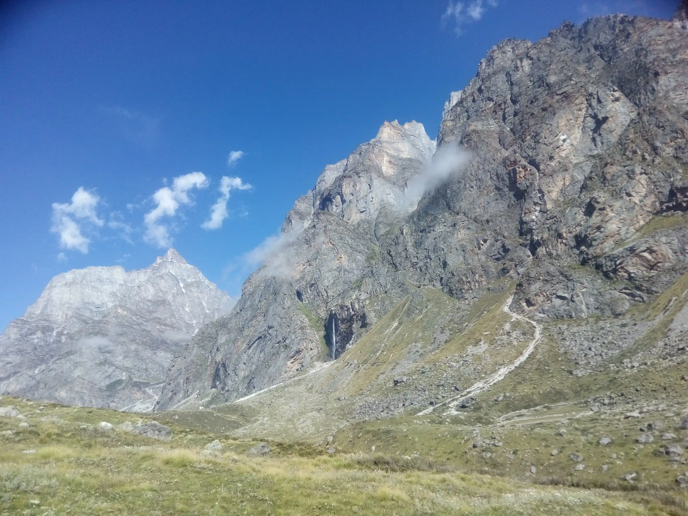 The stony trail climbing a green slope towards a thin white waterfall on a tall grey rock wall, with jagged peaks against a blue sky