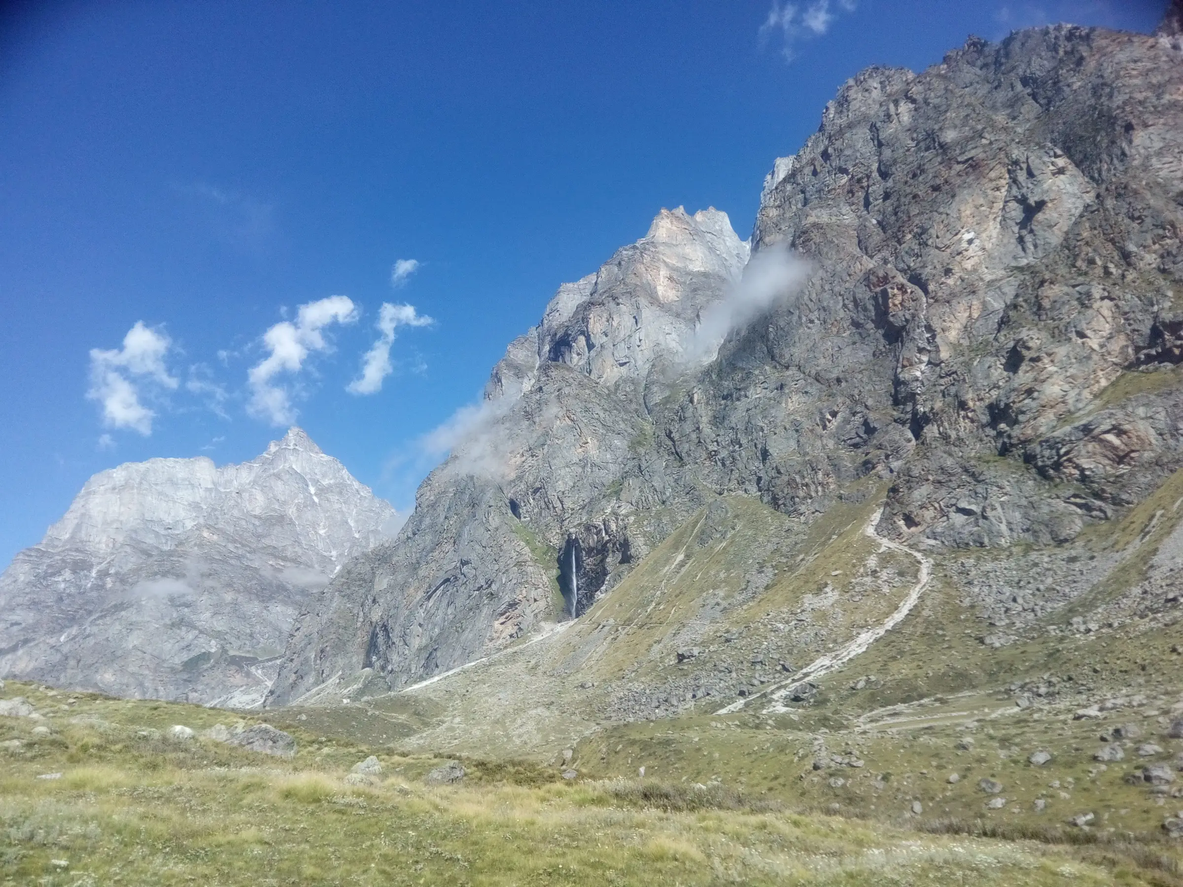 The stony trail climbing a green slope towards a thin white waterfall on a tall grey rock wall, with jagged peaks against a blue sky