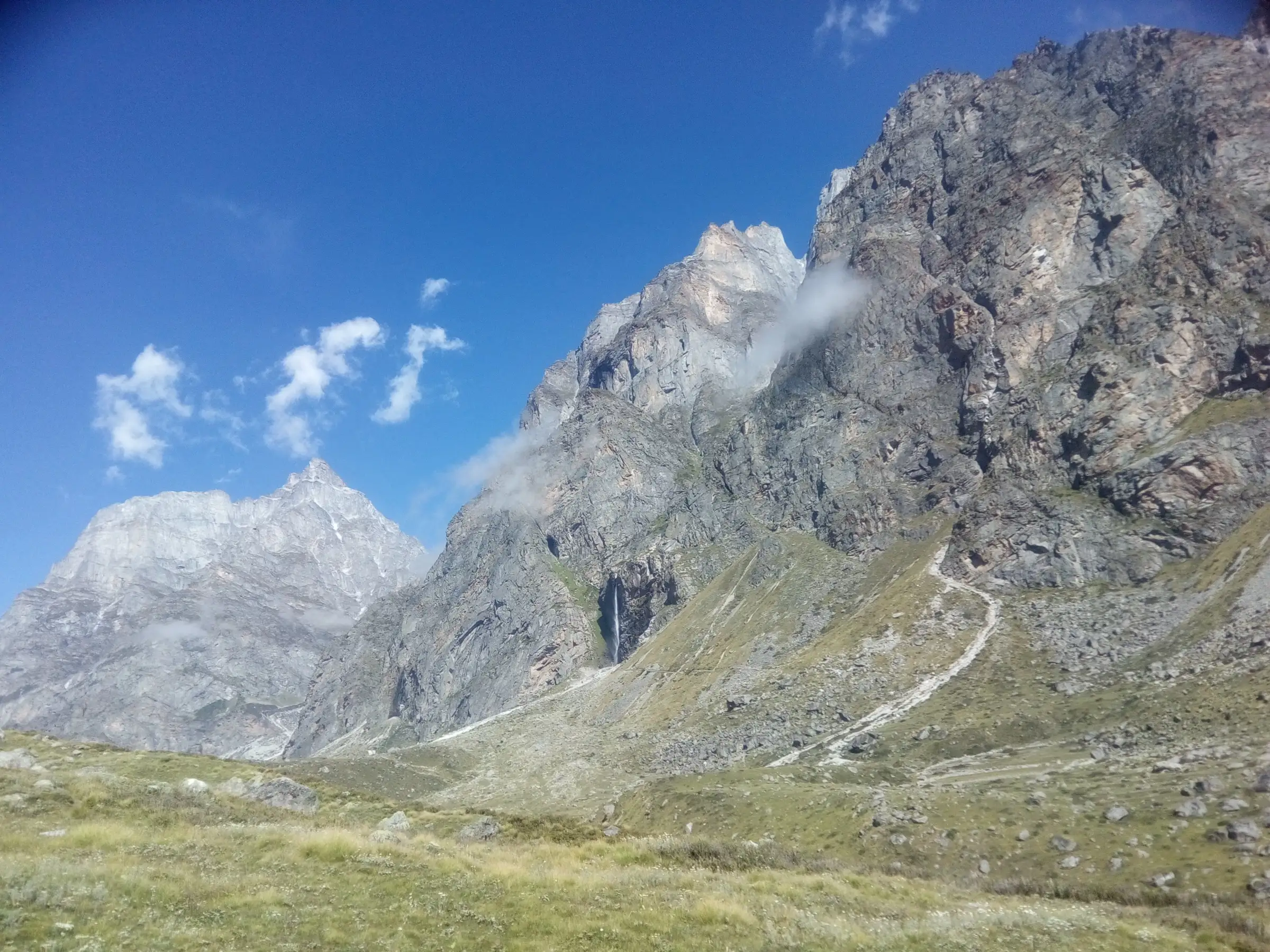 The stony trail climbing a green slope towards a thin white waterfall on a tall grey rock wall, with jagged peaks against a blue sky