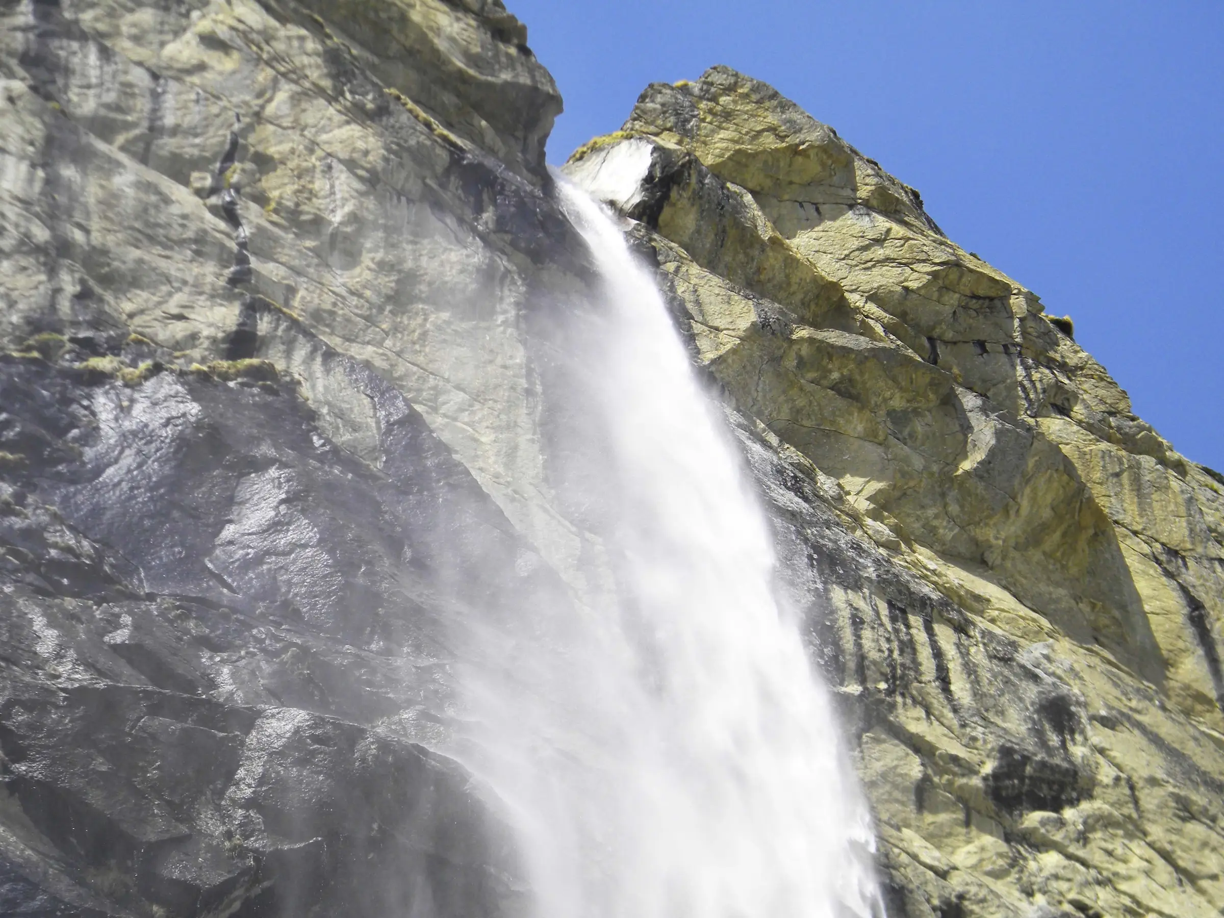 Looking up at Vasudhara Falls as it pours over a sunlit grey and yellow rock face into white spray under a clear blue sky