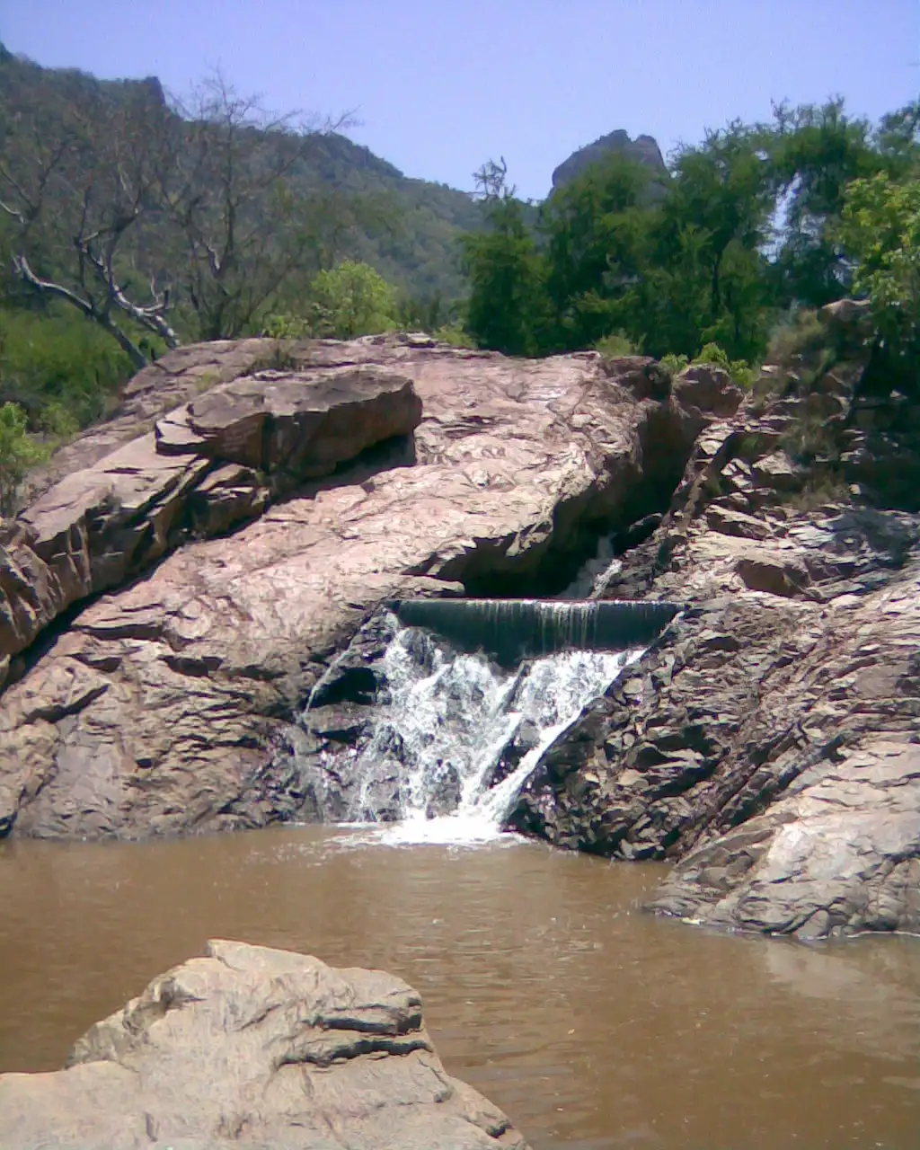 Water spilling over a small check-dam and pink granite slabs into a muddy pool in Amirthi forest