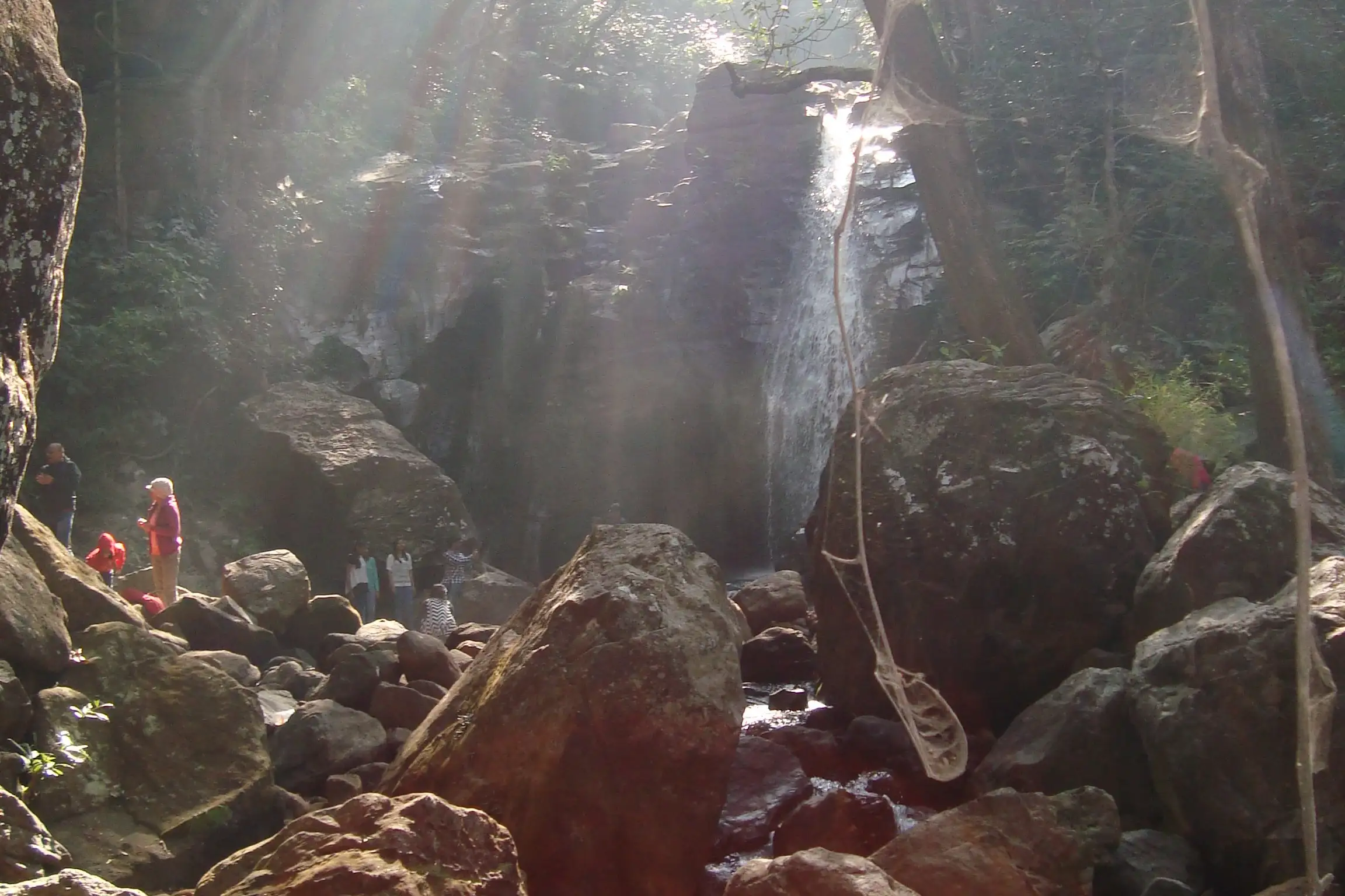 Sunlight streaming through forest onto Duchess Fall dropping over a dark rock face, with visitors standing among large boulders at its base