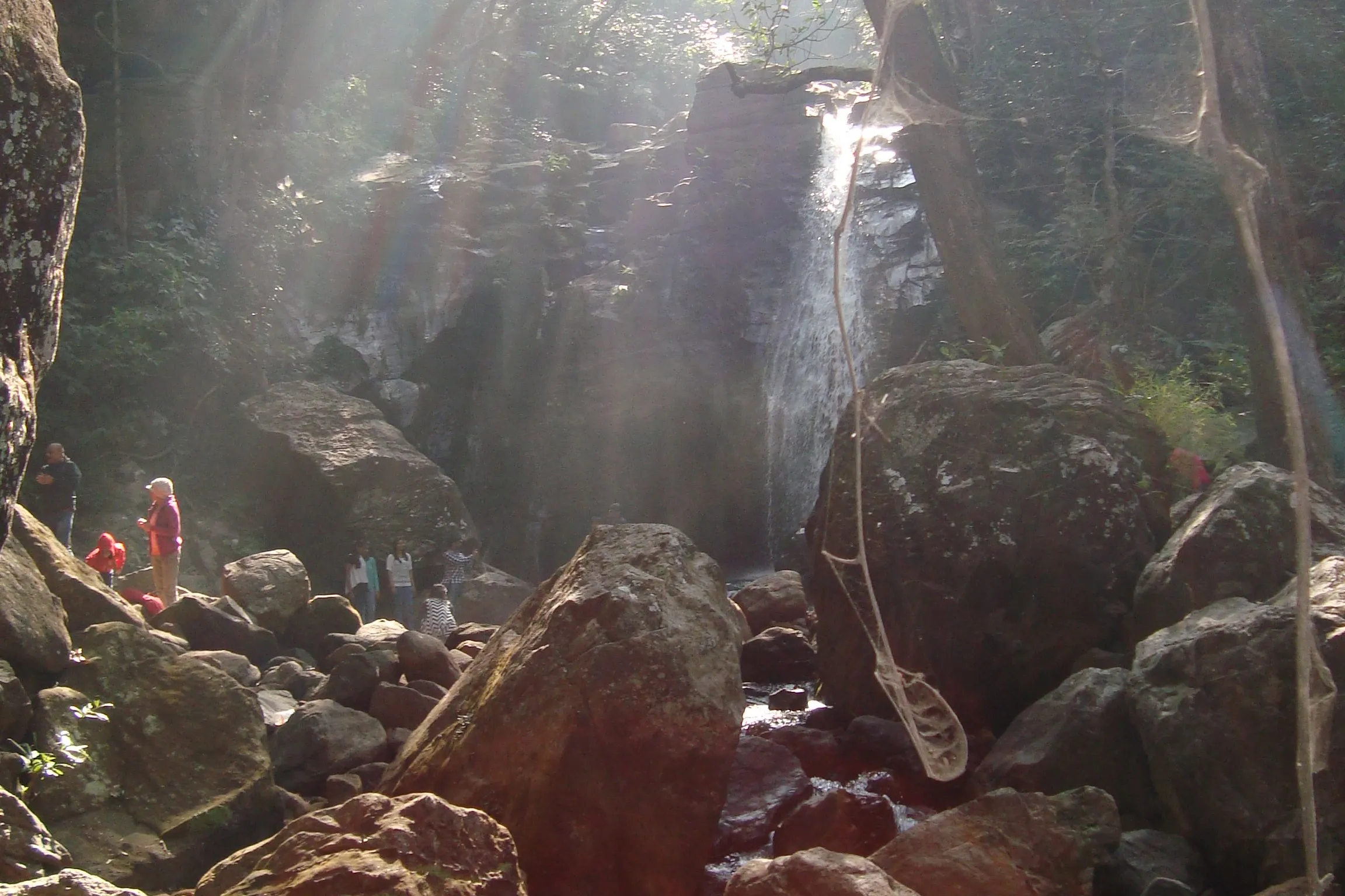 Sunlight streaming through forest onto Duchess Fall dropping over a dark rock face, with visitors standing among large boulders at its base