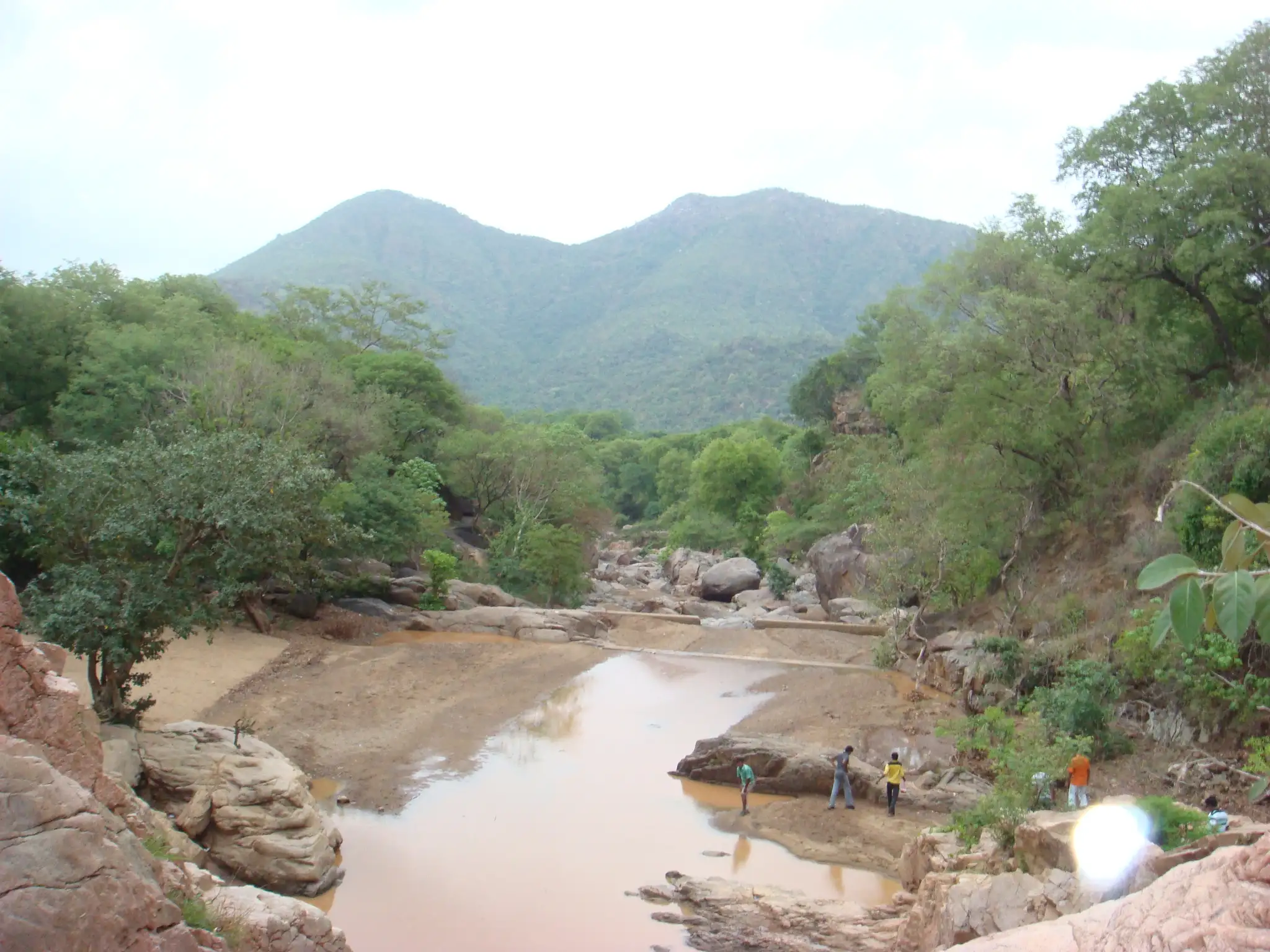 Rocky stream bed and pools in Amirthi Forest, with visitors on the rocks and forested hills behind