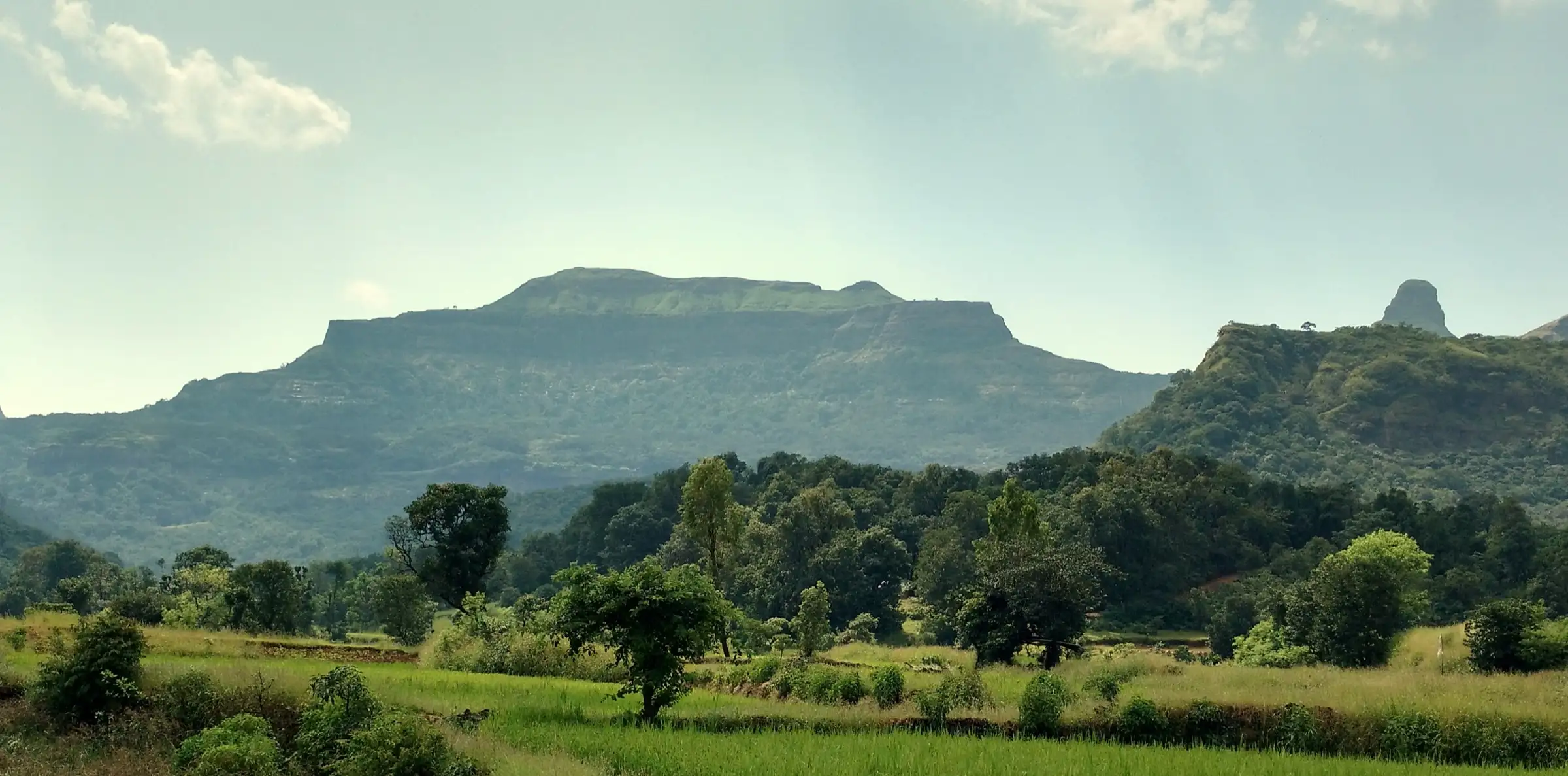 Flat-topped Ratangad fort rising above green fields and forest, with the thumb-shaped Khutta pinnacle on the right