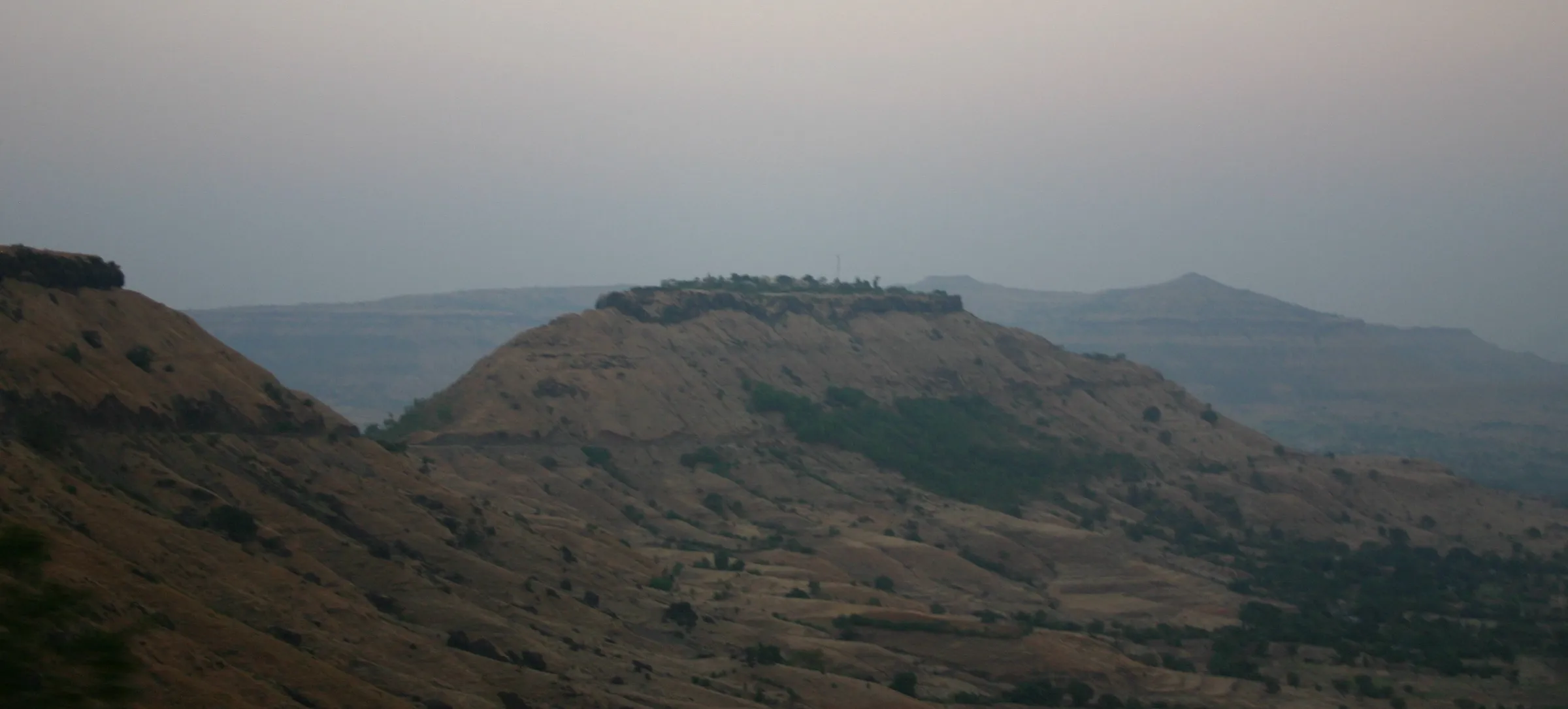 Flat-topped Sajjangad hill with its fort walls along the summit, seen across dry Sahyadri slopes in evening haze