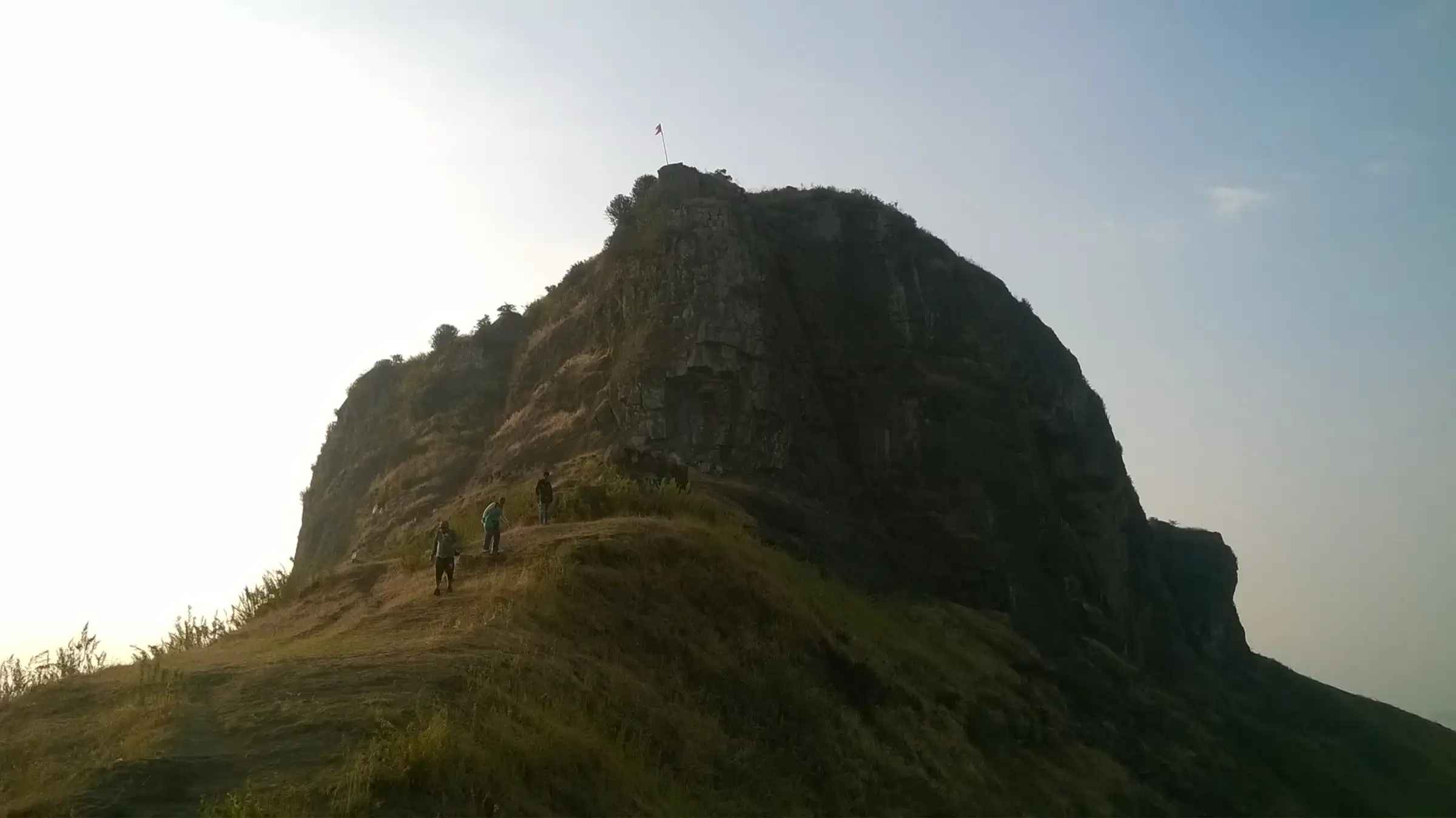 Trekkers on a grassy ridge below the rocky crown of Kavnai Fort, with a saffron flag on top