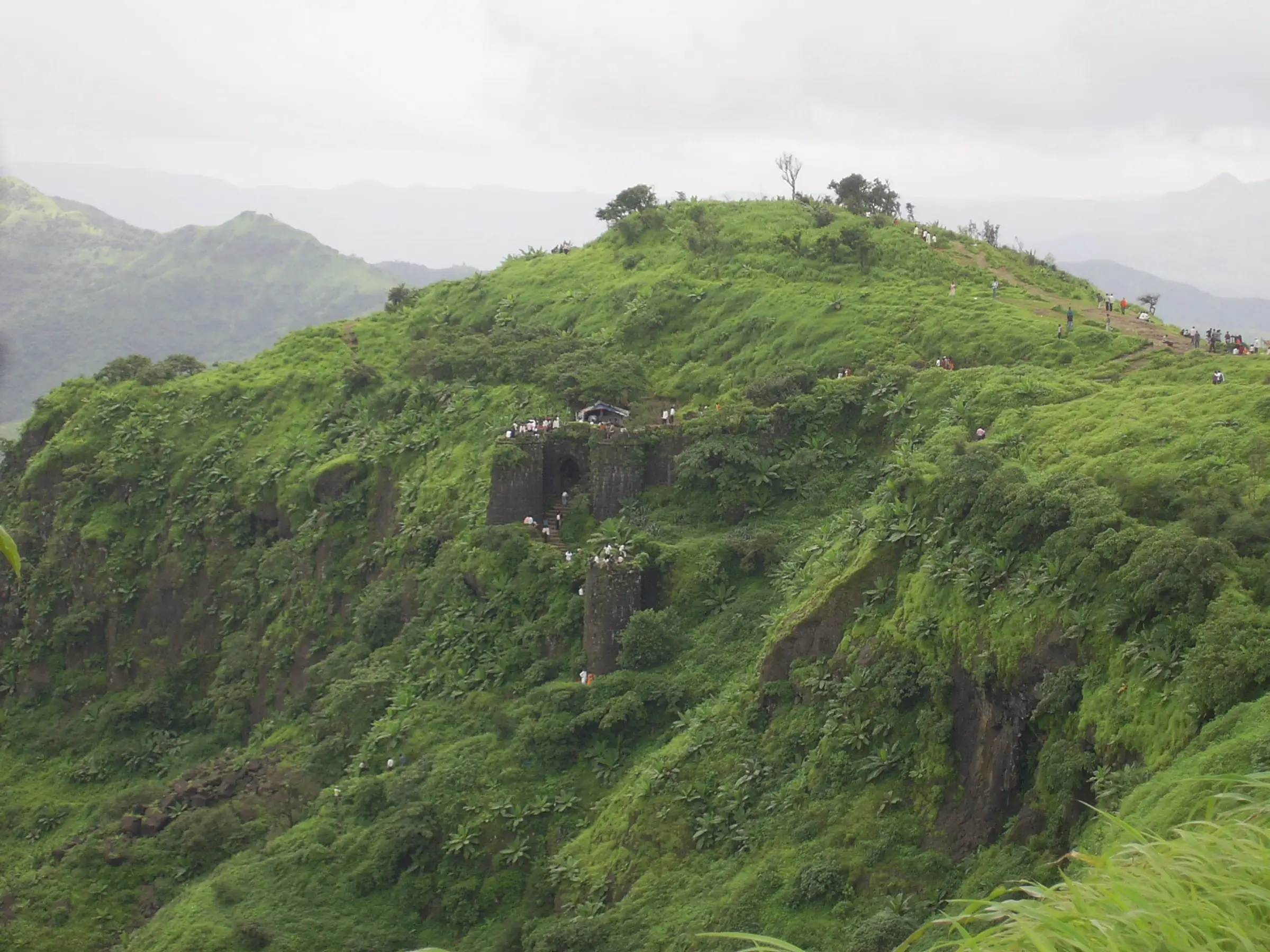 Green monsoon slopes of Sinhagad with old stone fort walls and a bastion on the hillside, walkers on the ridge above