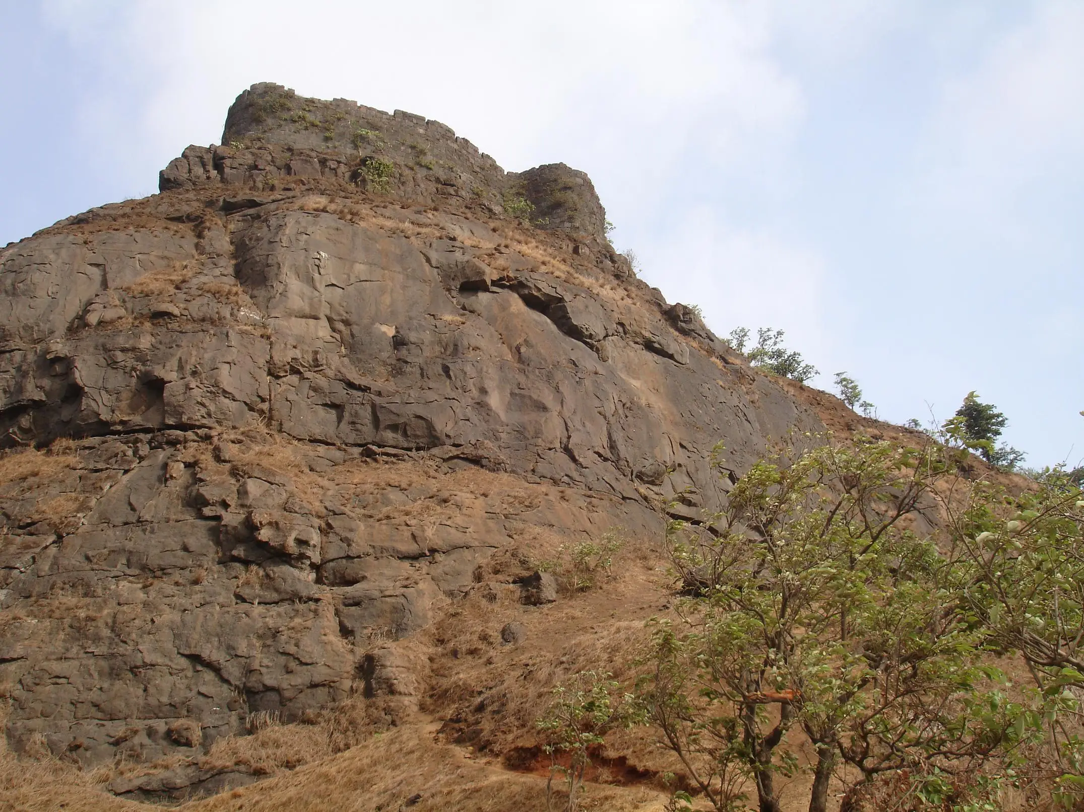 Rock scarp of Peb Fort (Vikatgad) rising above dry grass and trees, with the top ramparts on the skyline