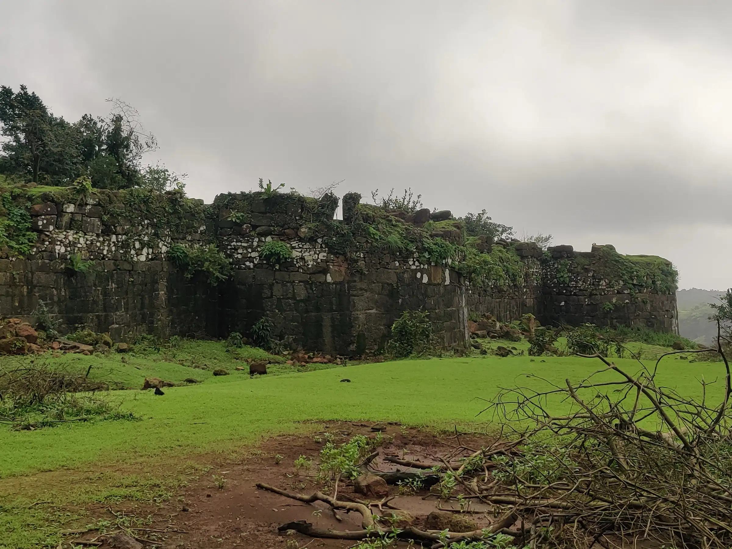 Long ruined stone rampart of Sagargad fort behind a green meadow under grey monsoon clouds