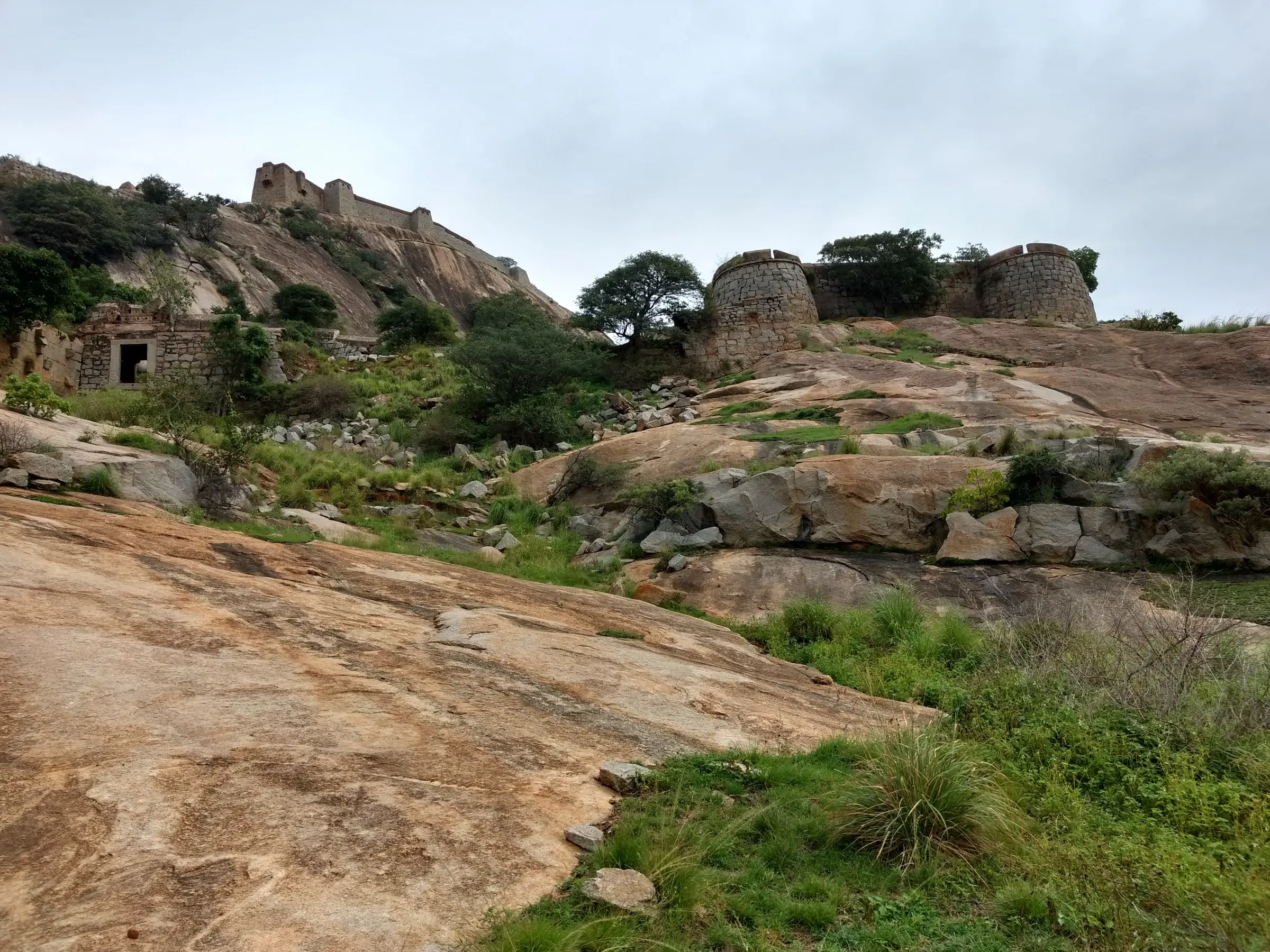 Granite slabs and green scrub leading up to round stone bastions and ruined fort walls on Channarayana Durga