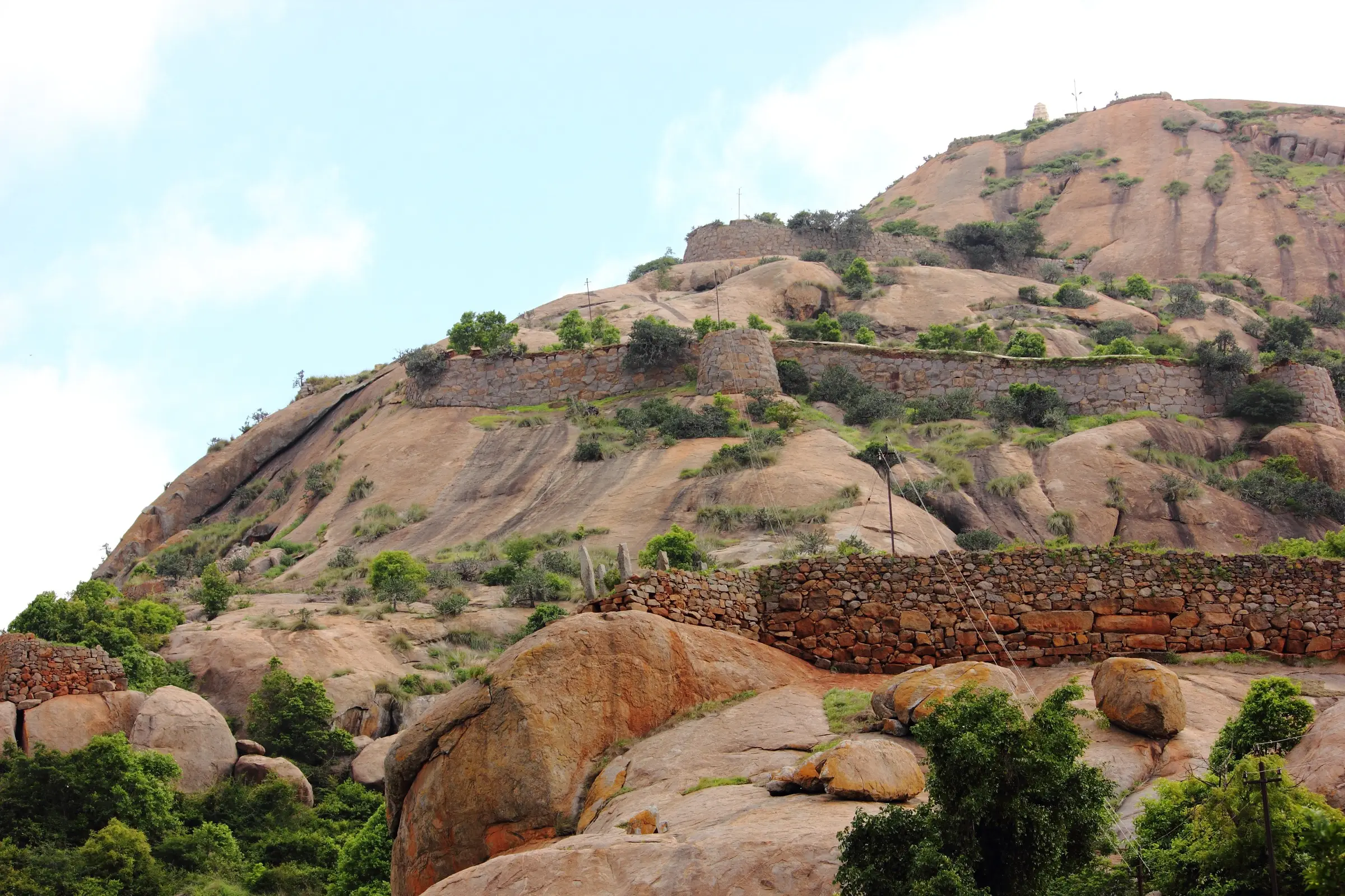 Old stone fort walls climbing a granite hill at Hutridurga, with boulders and green scrub in front