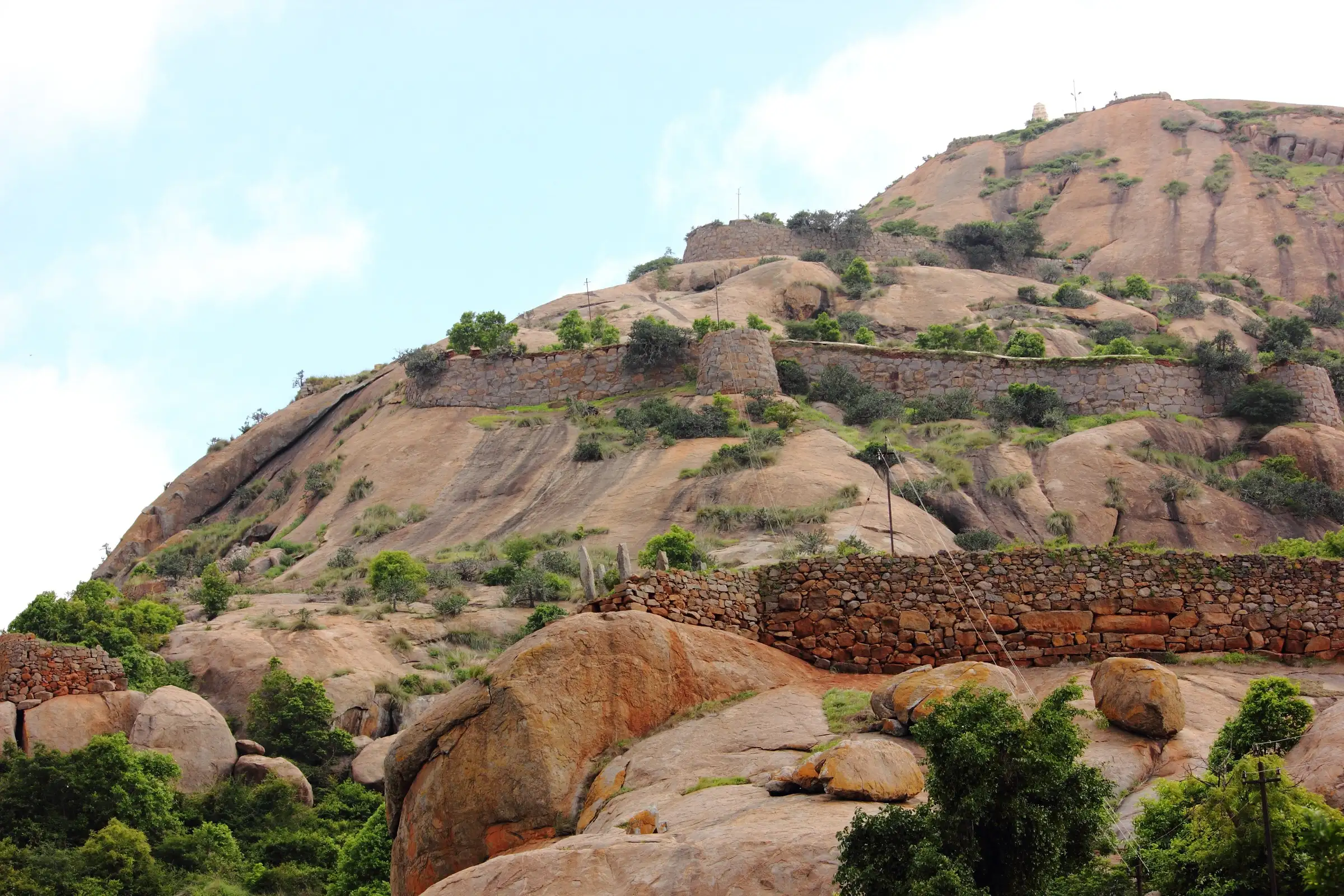 Old stone fort walls climbing a granite hill at Hutridurga, with boulders and green scrub in front