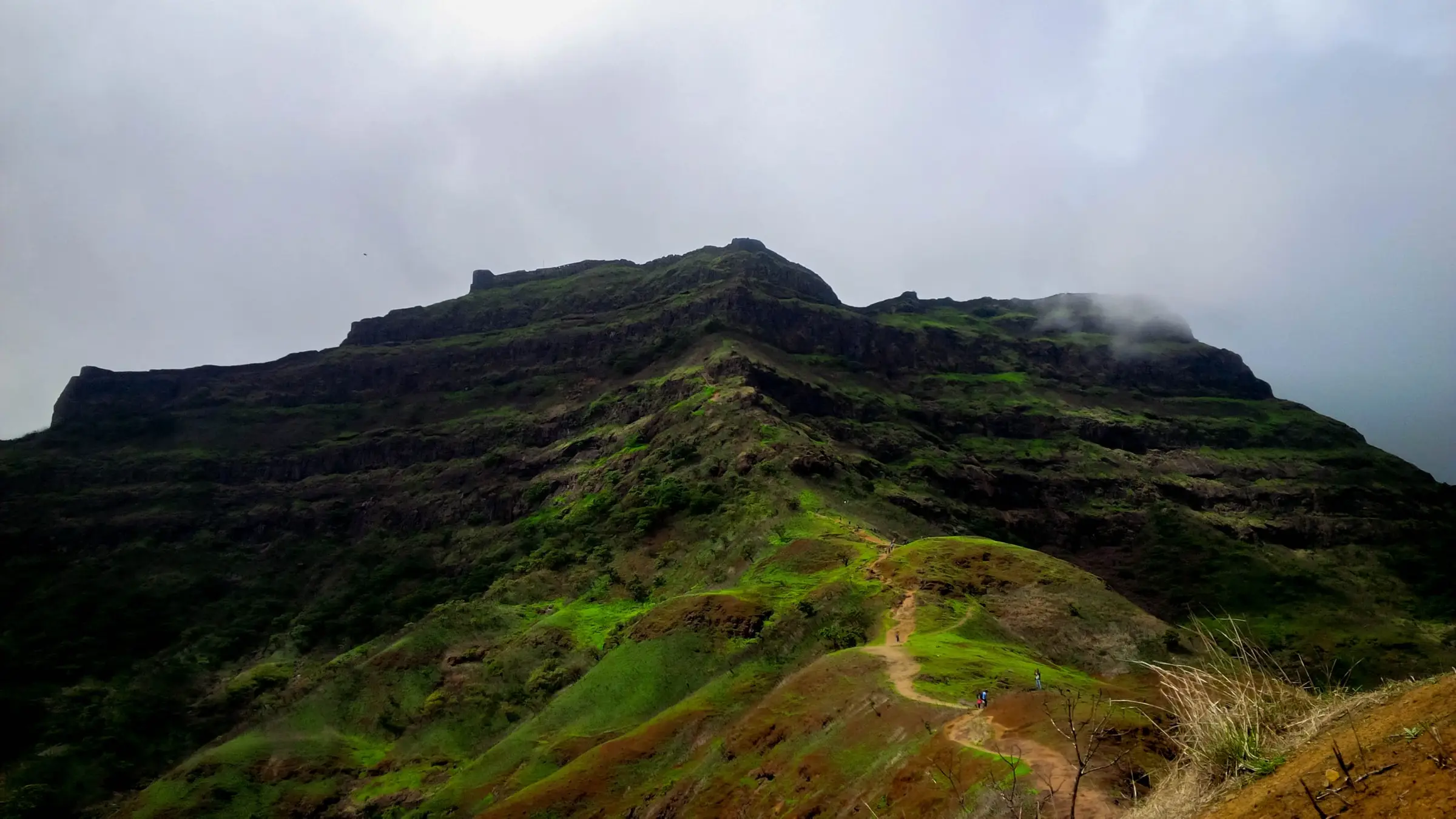 The long flat-topped ridge of Torna Fort under monsoon cloud, with a dirt trail climbing a green spur towards it