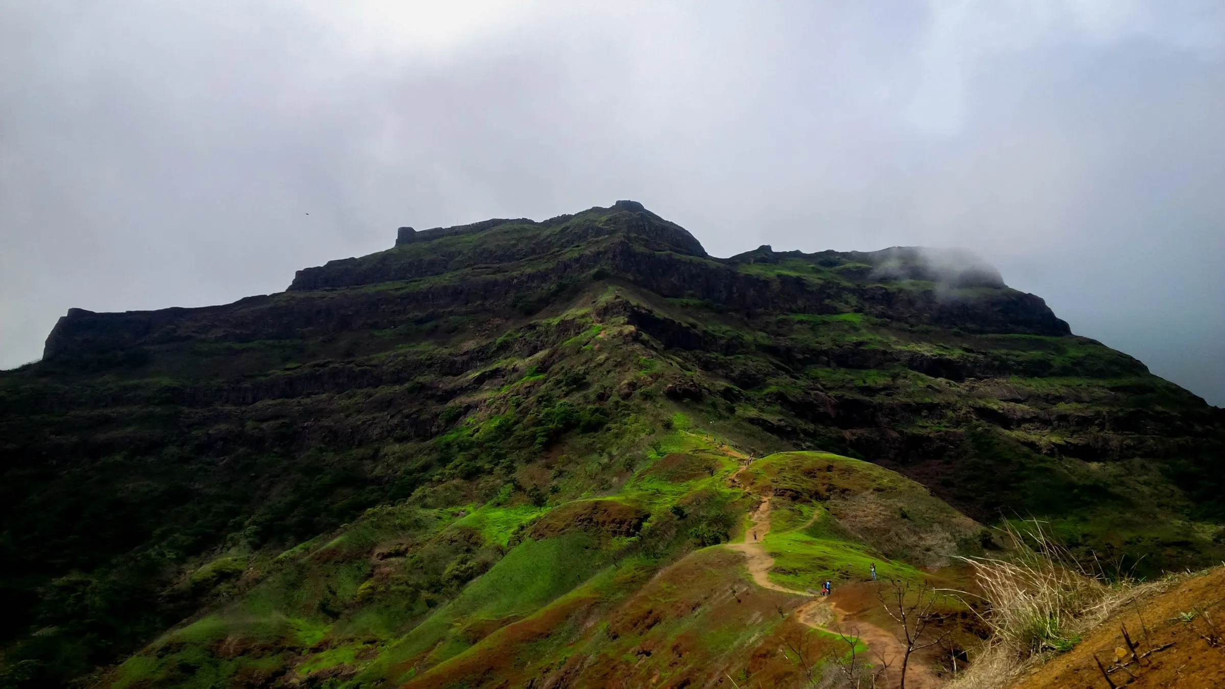 The long flat-topped ridge of Torna Fort under monsoon cloud, with a dirt trail climbing a green spur towards it