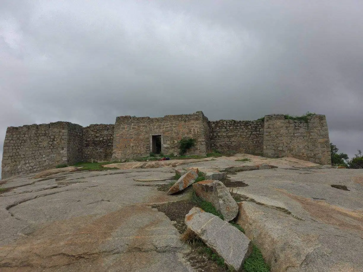 Stone walls and a doorway of the Medigeshi Fort ruins on the bare granite summit under grey monsoon clouds