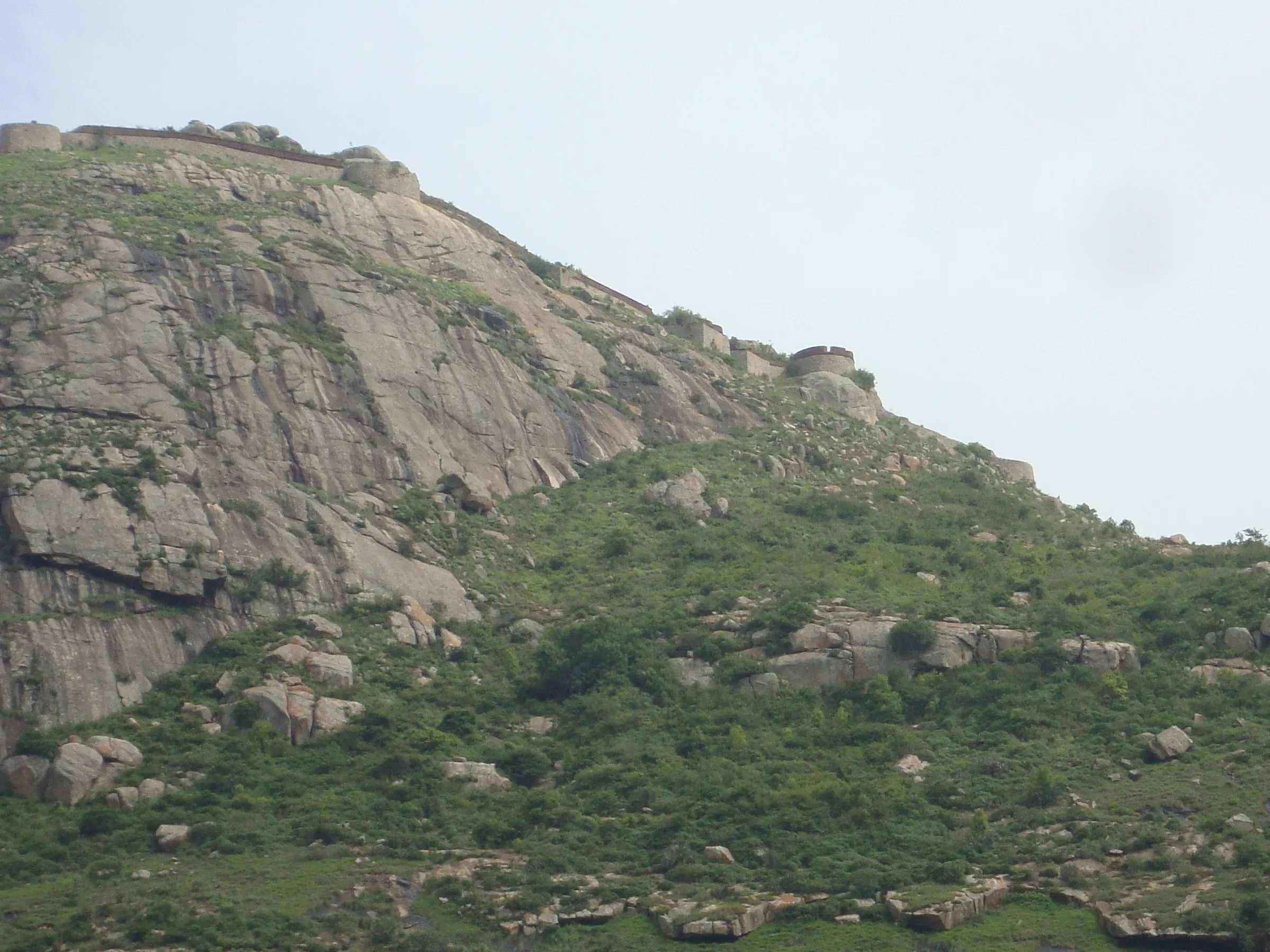 Rocky granite face of Kaiwara Betta with green scrub below and the fort walls and bastions along the summit ridge