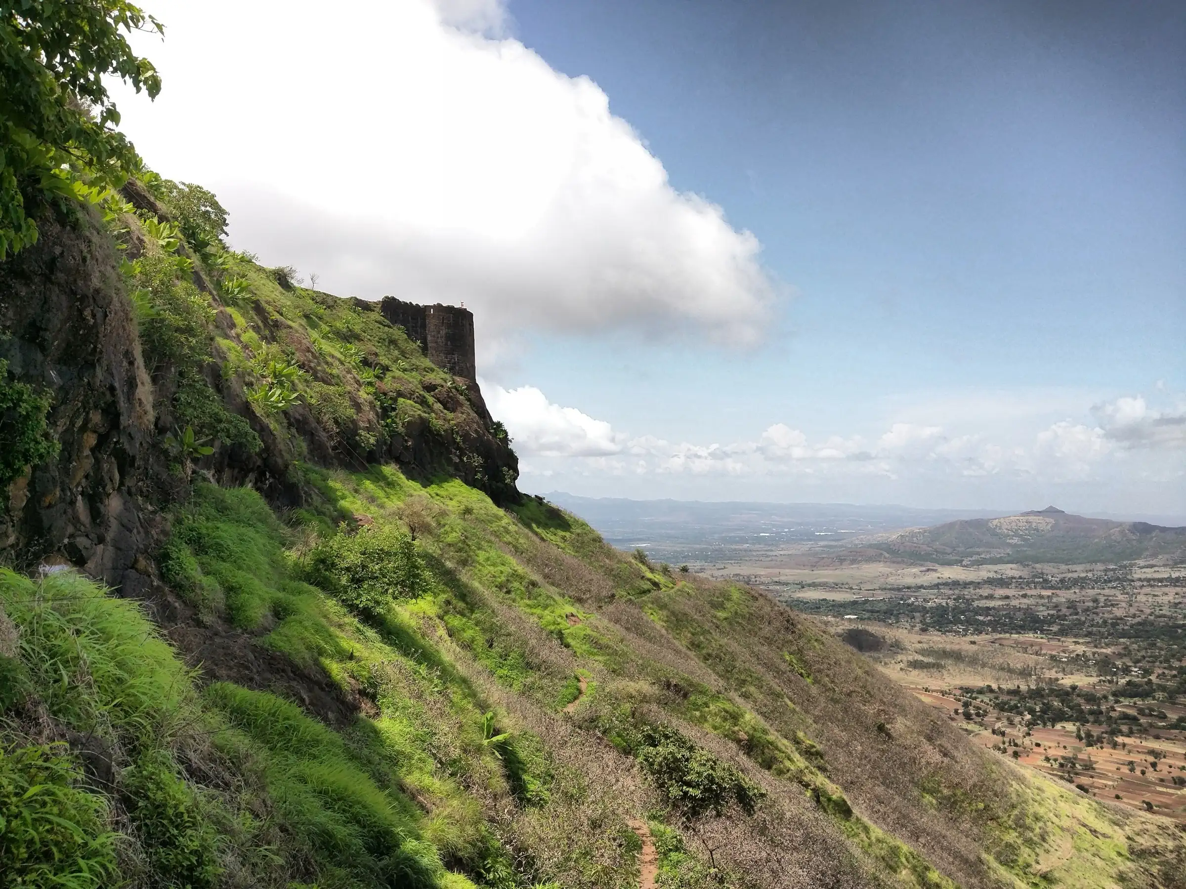 A stone bastion of Rohida fort on a green slope above the wide plain and hills, seen from the first gateway