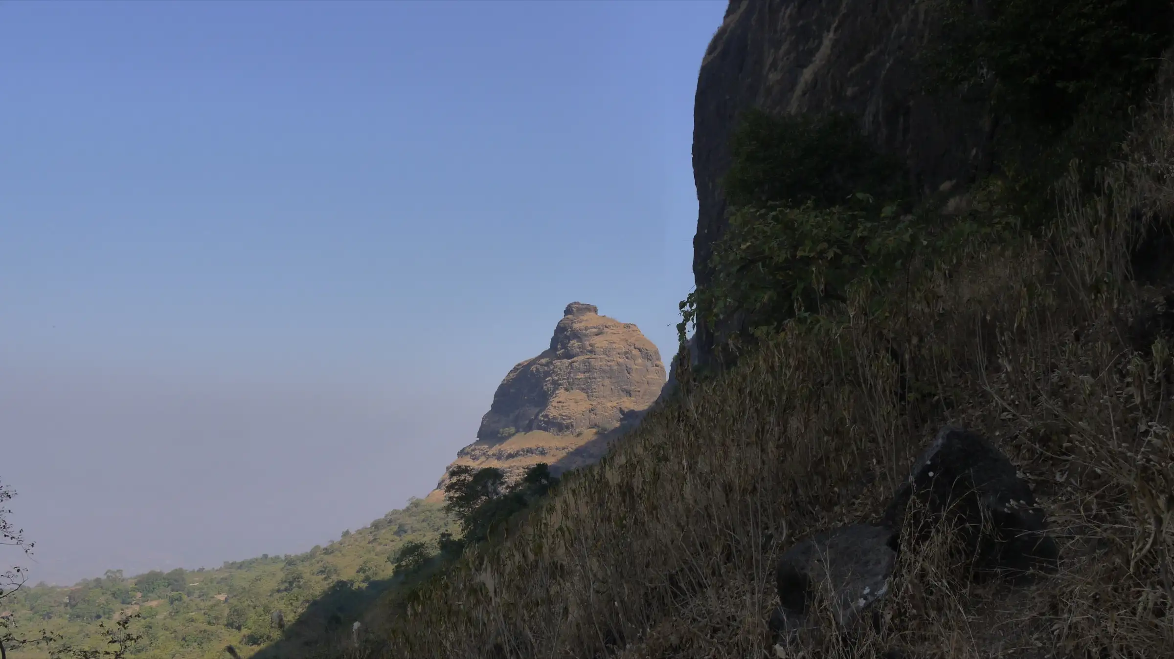 Kalavantin Durg rising beyond a dry grassy slope, seen from Prabal Machi in winter haze