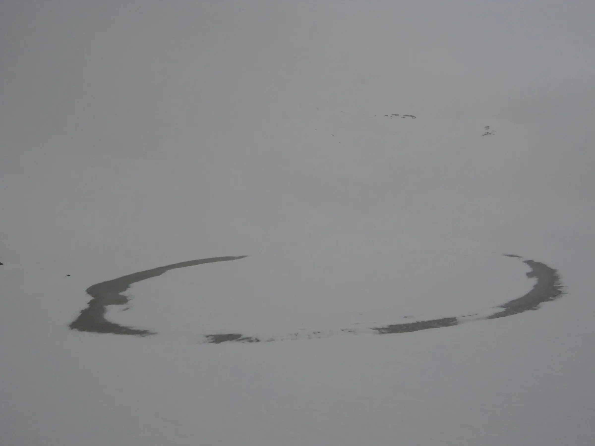Frozen Bhrigu Lake in mist, with only a thin dark ring of open water showing around its snow-covered surface