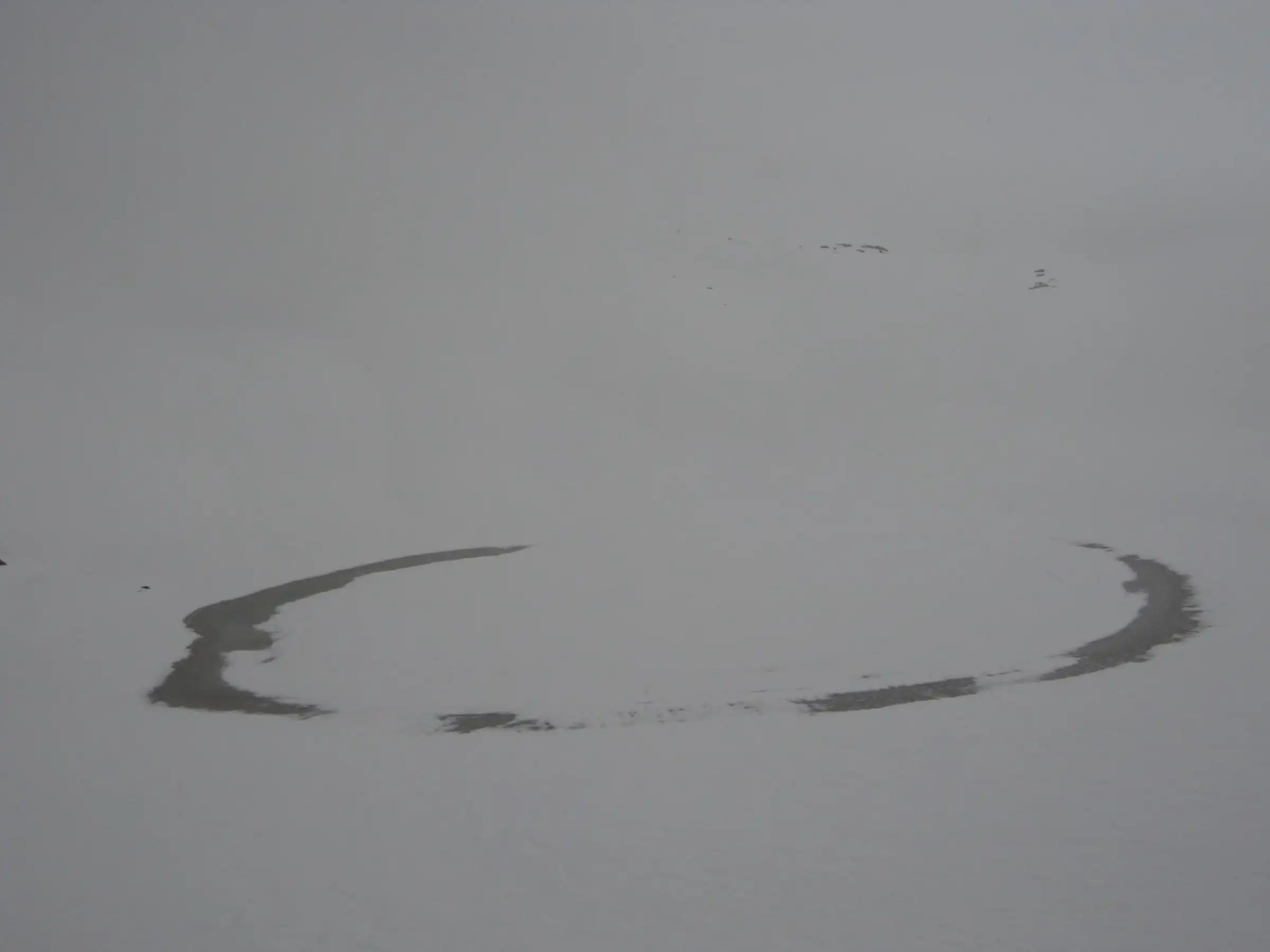 Frozen Bhrigu Lake in mist, with only a thin dark ring of open water showing around its snow-covered surface