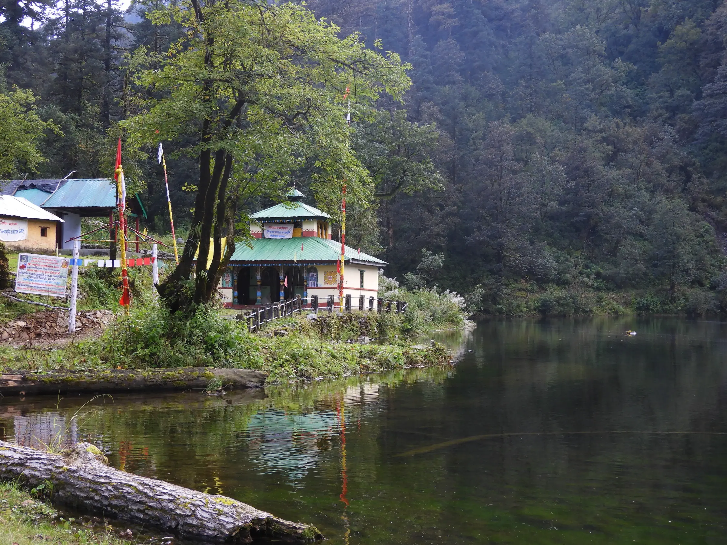 The green-roofed Shri Ganesh temple with prayer flags on the forested shore of Dodital lake, reflected in the water