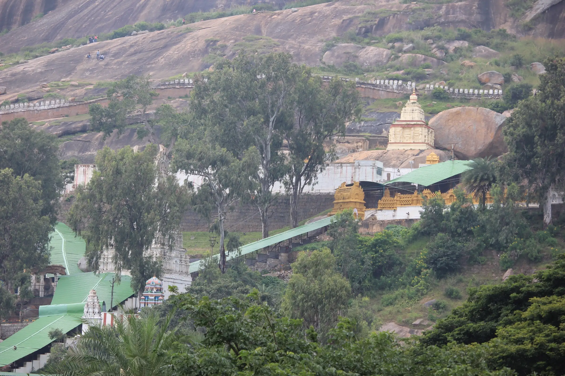 The Gavi Gangadhareshwara temple complex with green-roofed walkways and a white vimana against a huge boulder at the foot of Shivagange hill