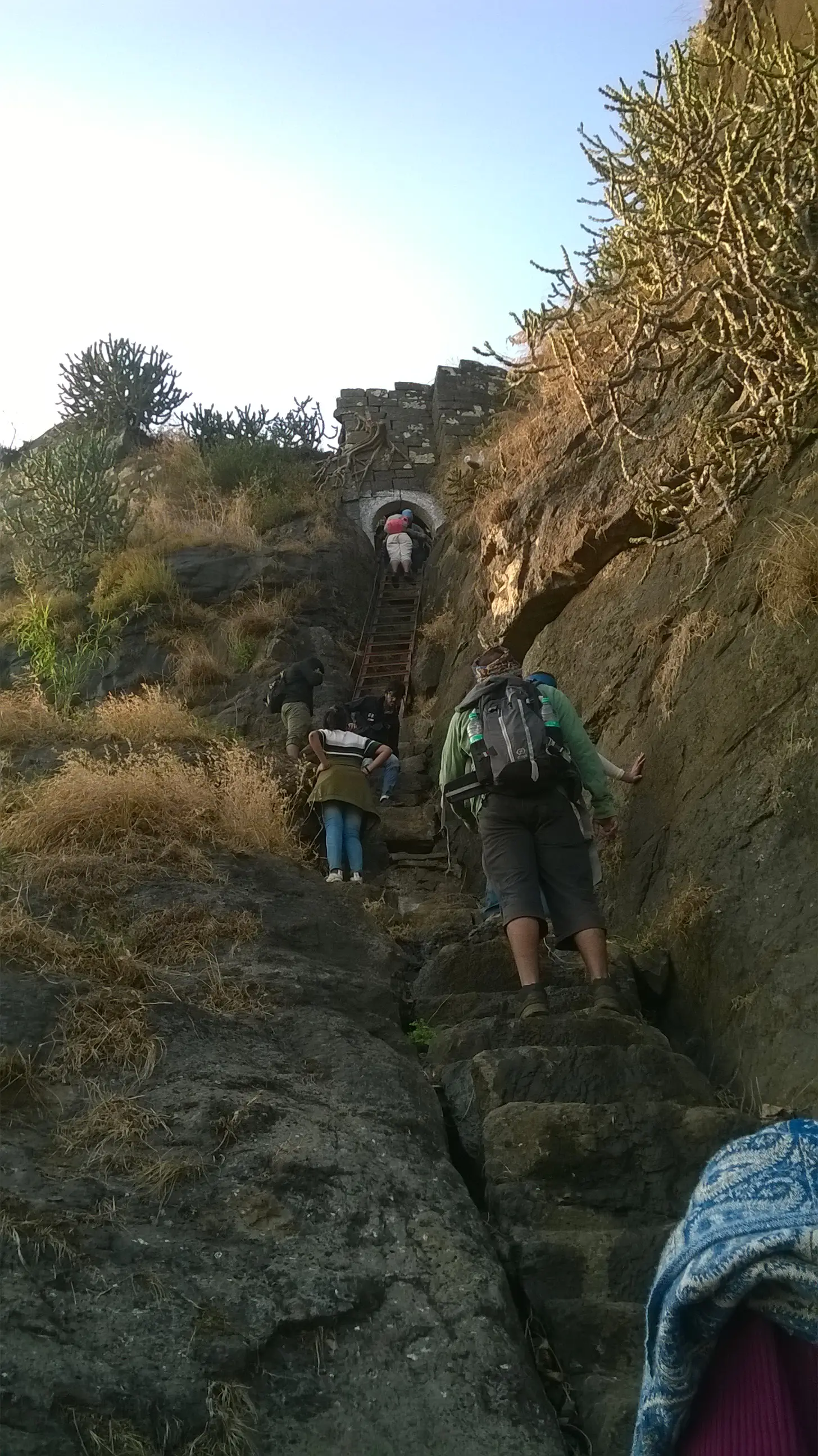 Trekkers climbing worn rock-cut steps and an iron ladder in a narrow gully up to the arched gate of Kavnai Fort