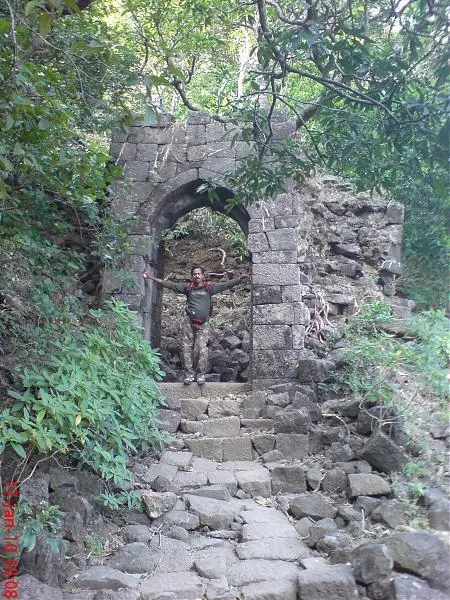 Ruined stone arched gateway on Siddhagad with rock-cut steps leading up through forest and a trekker standing in the arch