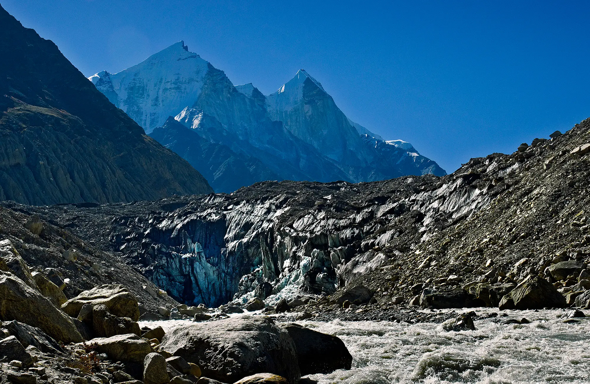 The grey ice face of the Gangotri Glacier snout at Gaumukh, with the Bhagirathi river rushing out over boulders and the snow-covered Bhagirathi peaks behind