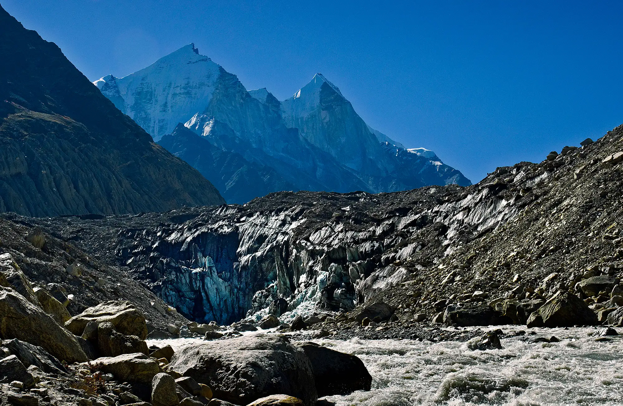 The grey ice face of the Gangotri Glacier snout at Gaumukh, with the Bhagirathi river rushing out over boulders and the snow-covered Bhagirathi peaks behind