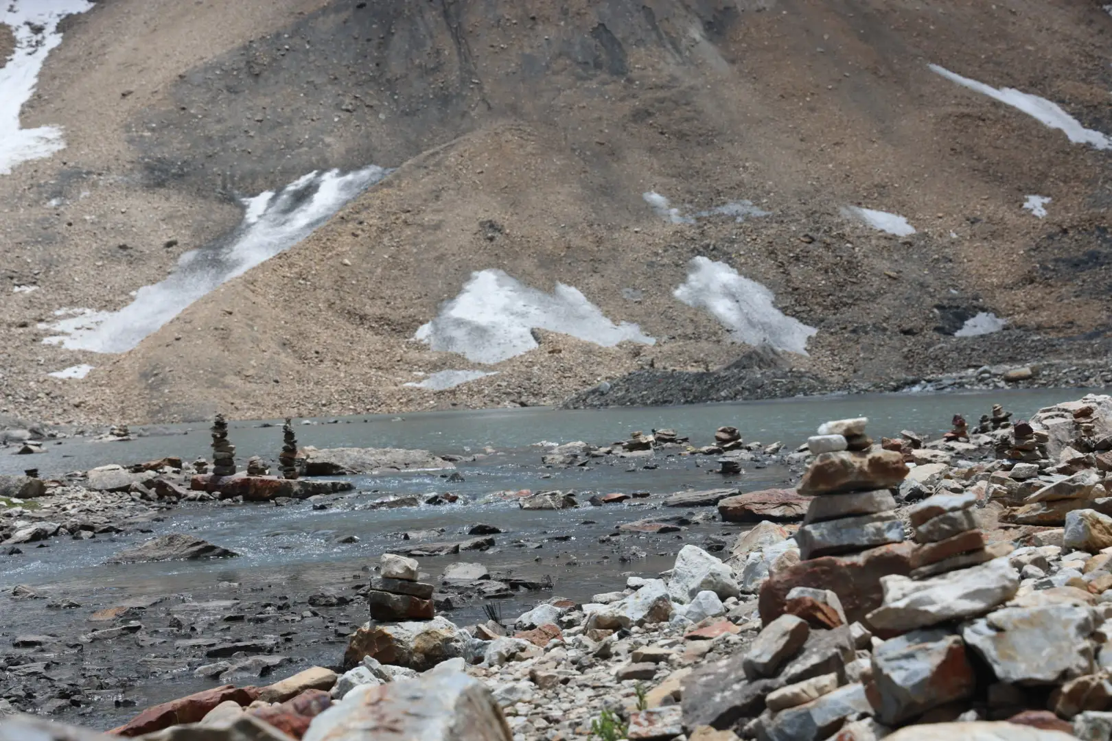 Grey glacial lake of Gauri Kund near Jolingkong ringed by stone cairns below bare scree slopes with snow patches