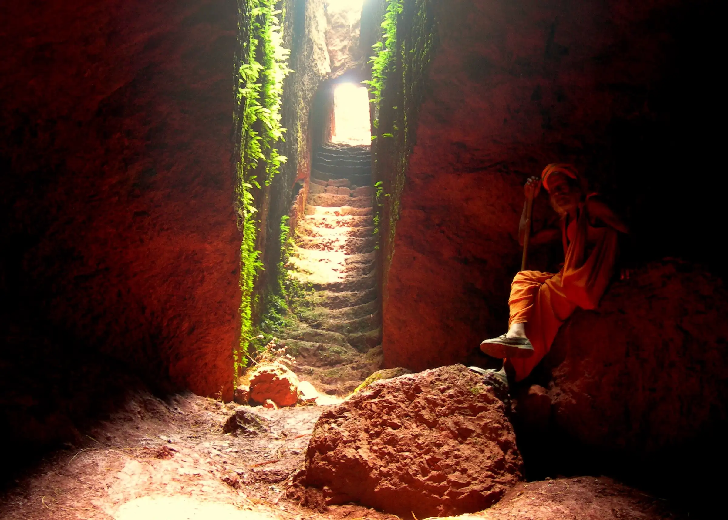 Inside the red-ochre rock well on Kamalgad, with steps rising to daylight and a man in saffron seated on a rock