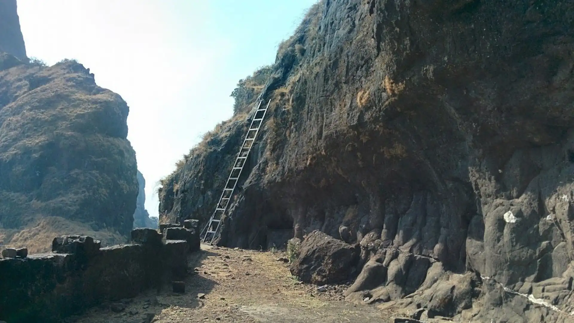 The steel ladder leaning against the rock face of Ghangad Fort above a level path lined with a low stone wall