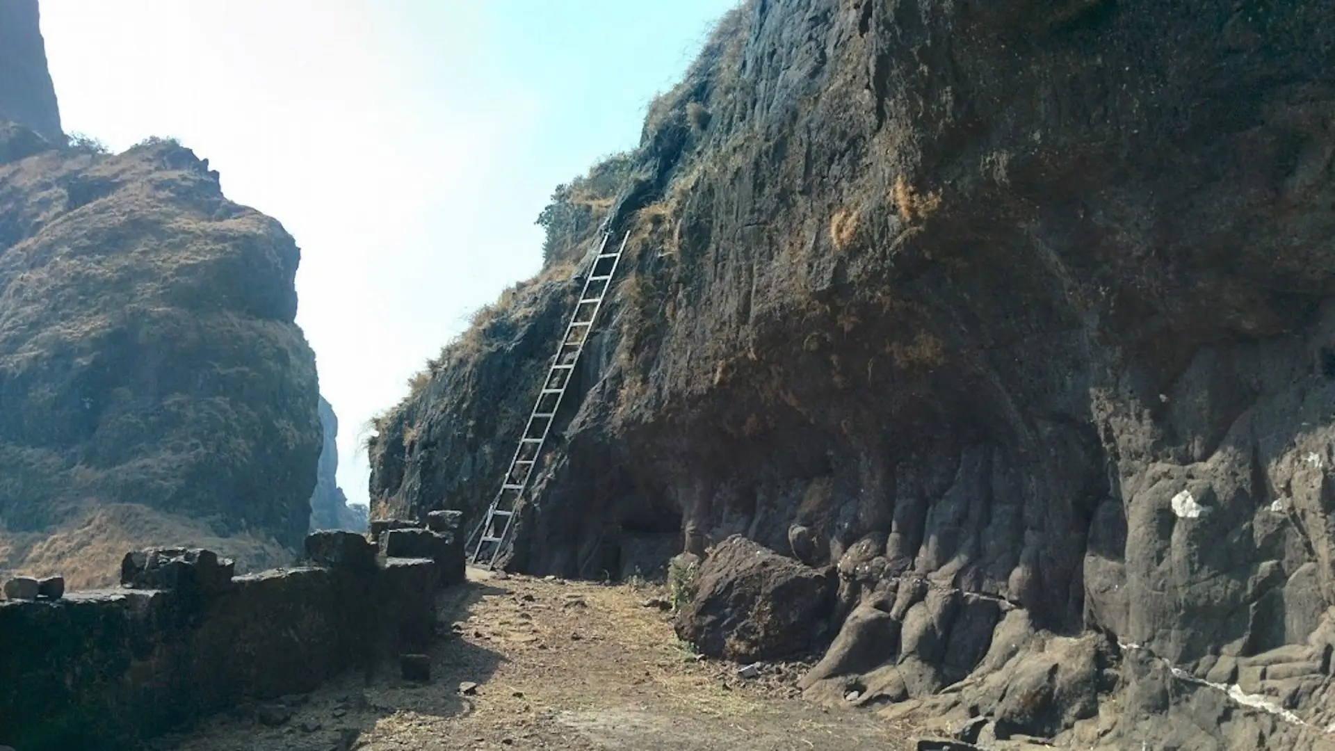 The steel ladder leaning against the rock face of Ghangad Fort above a level path lined with a low stone wall