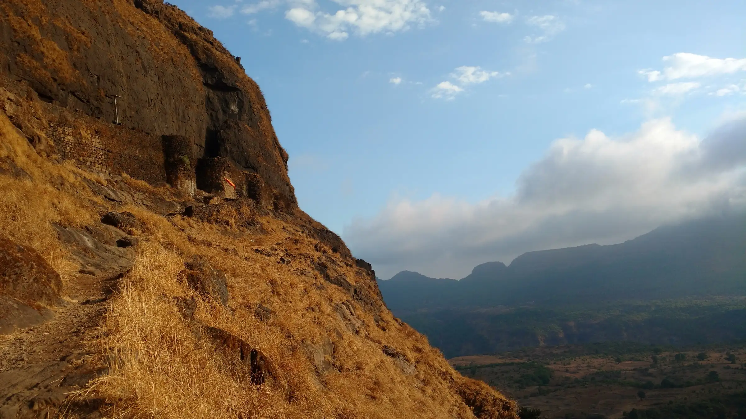 A narrow path through dry golden grass below the Ghangad scarp and its fortification walls, with Sahyadri ridges beyond