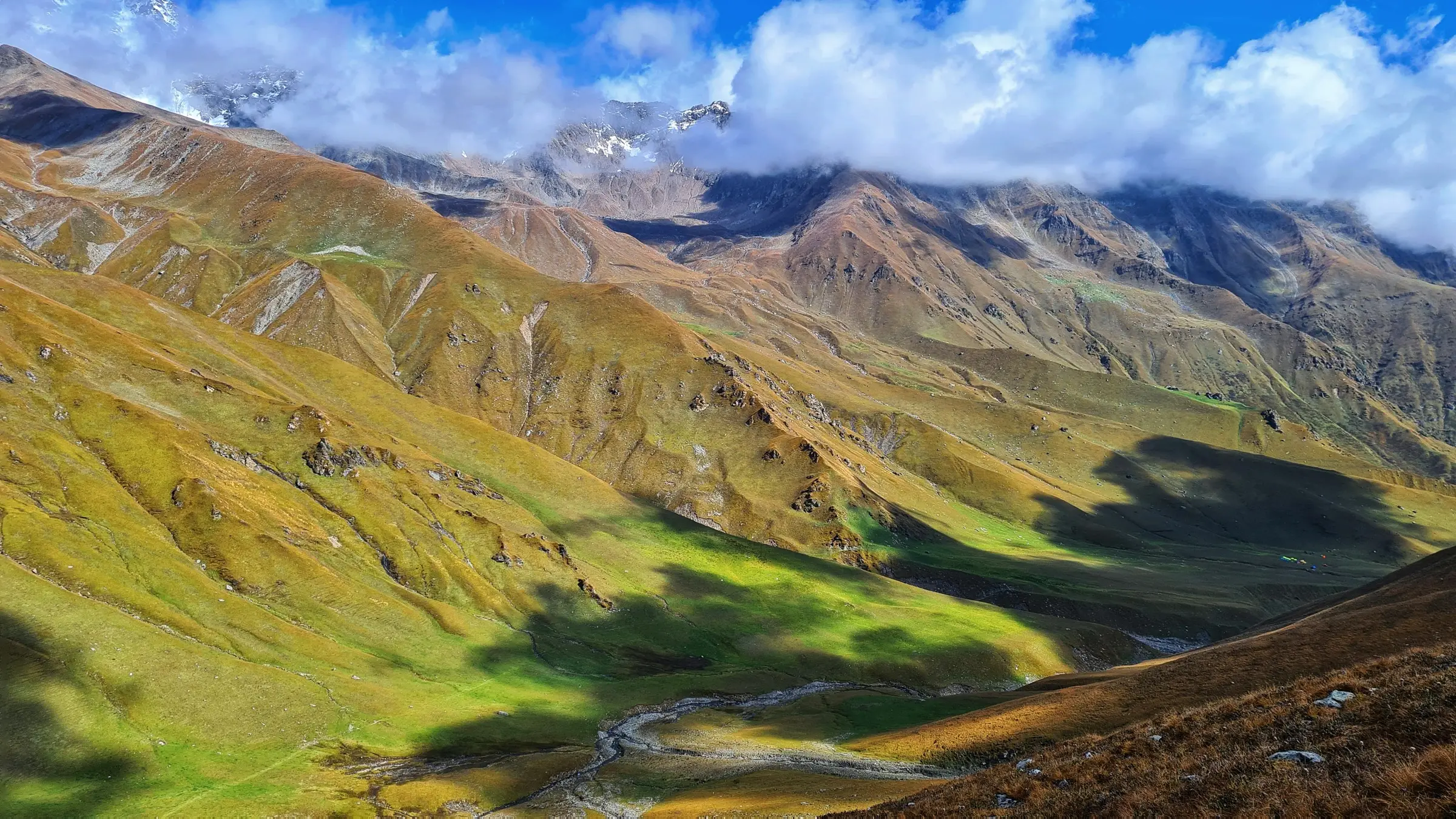 Green and golden Gidara Bugyal meadows falling to a stream, with snow peaks half hidden in cloud above