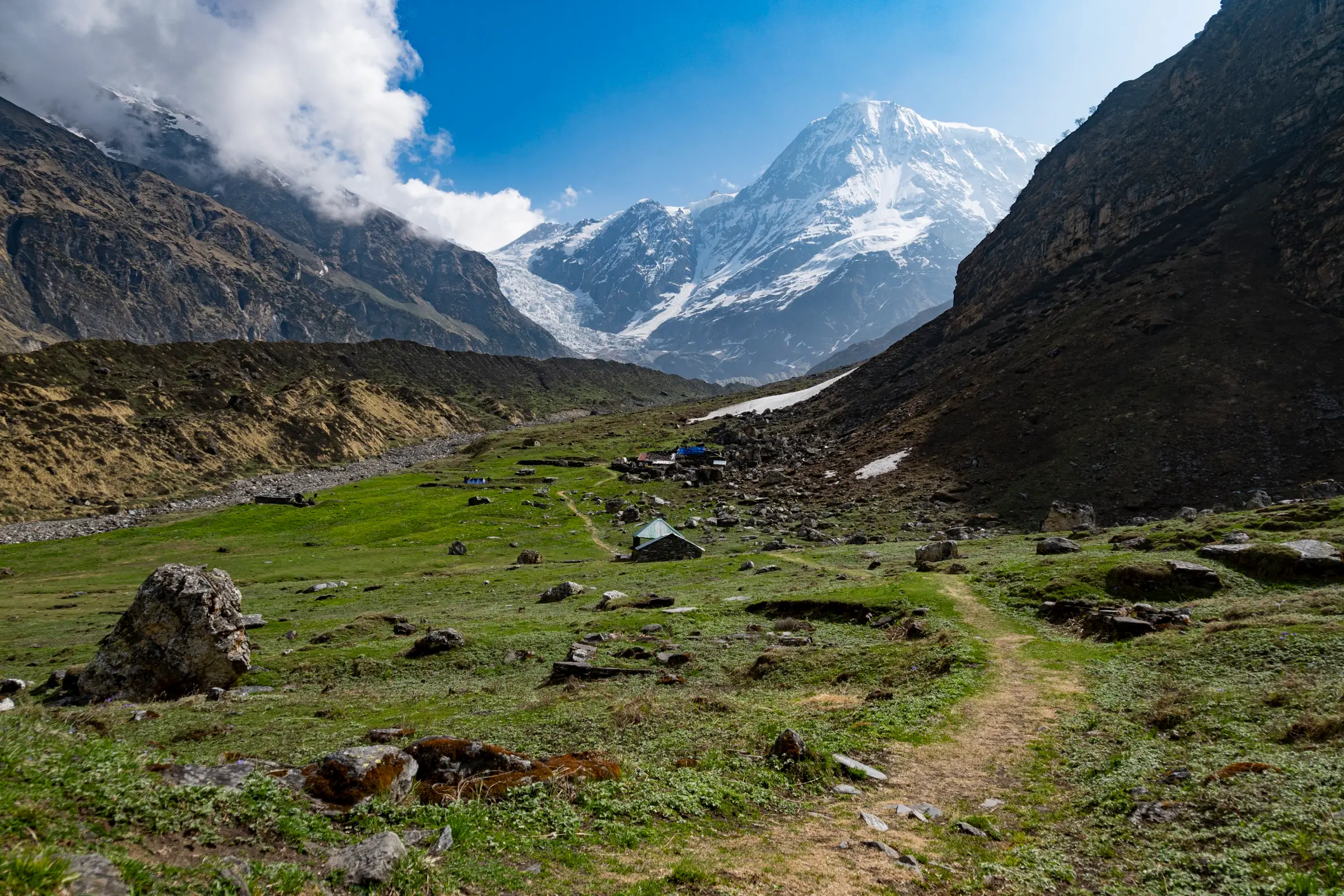 Green meadow with boulders and a few huts on the trail towards Pindari Glacier, a snowy peak at the head of the valley
