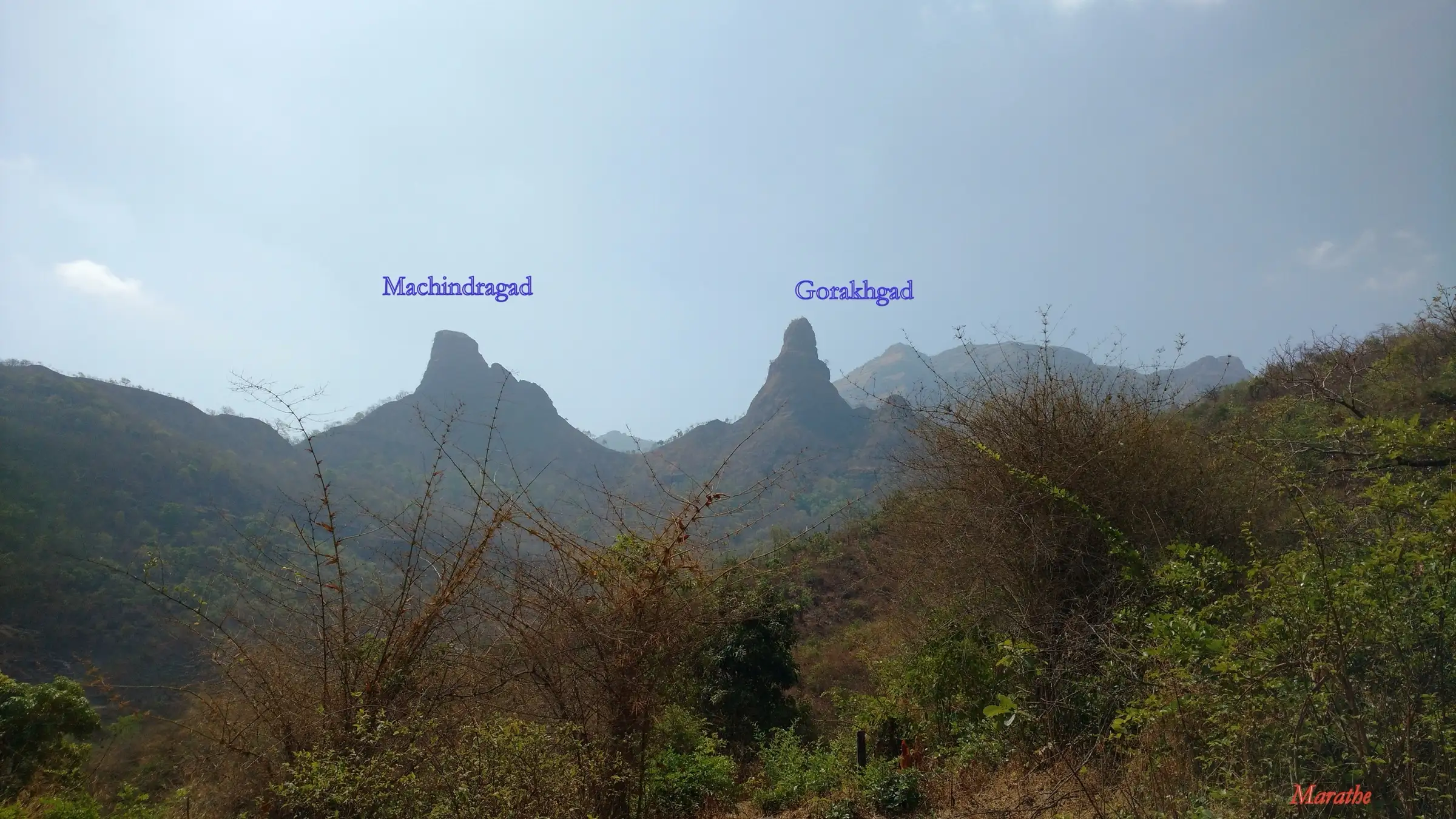 The twin rock pinnacles of Machindragad and Gorakhgad rising above dry forested hills, labelled with their names