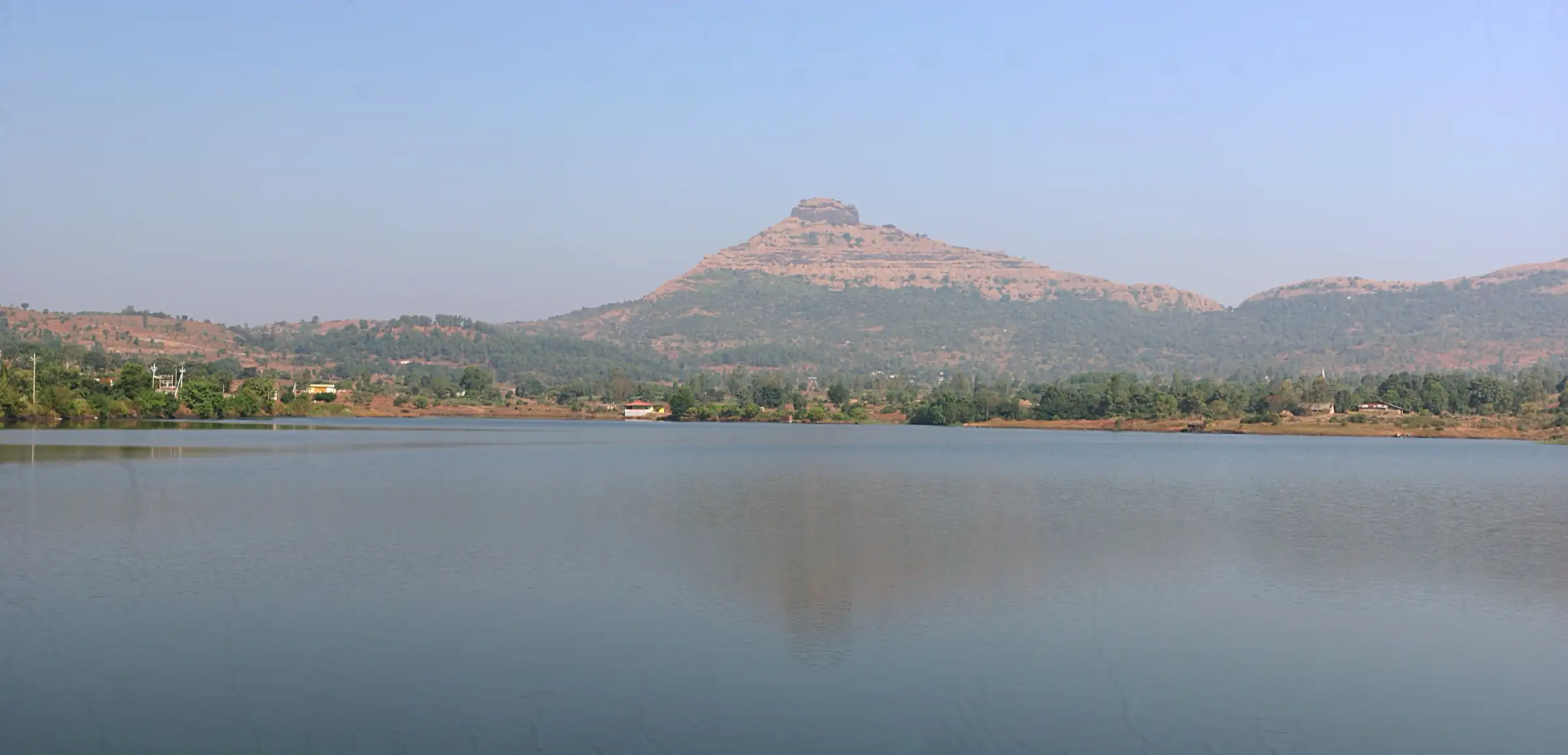 The pyramid-shaped Tikona Fort hill seen across the still water of Hadashi dam under a clear sky