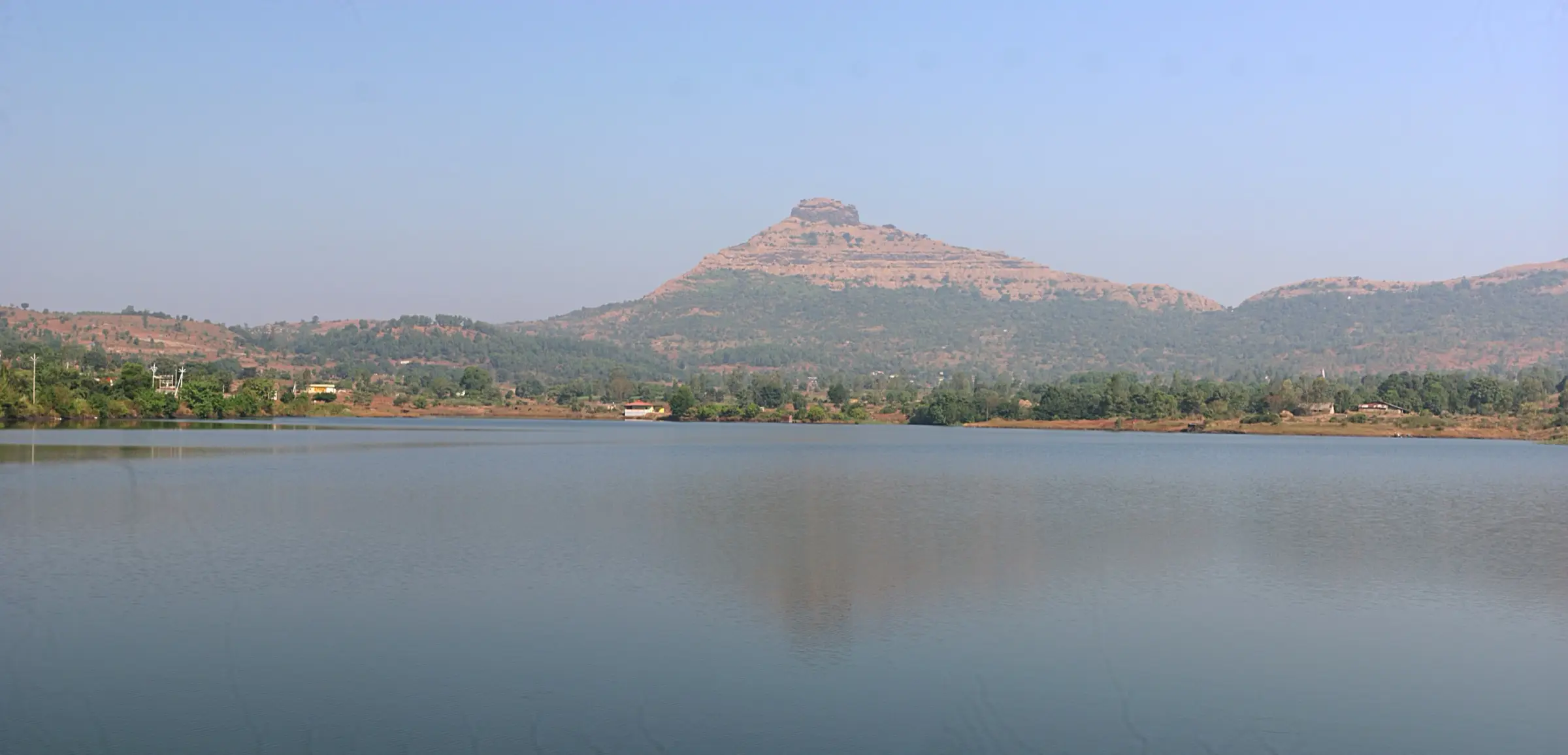 The pyramid-shaped Tikona Fort hill seen across the still water of Hadashi dam under a clear sky